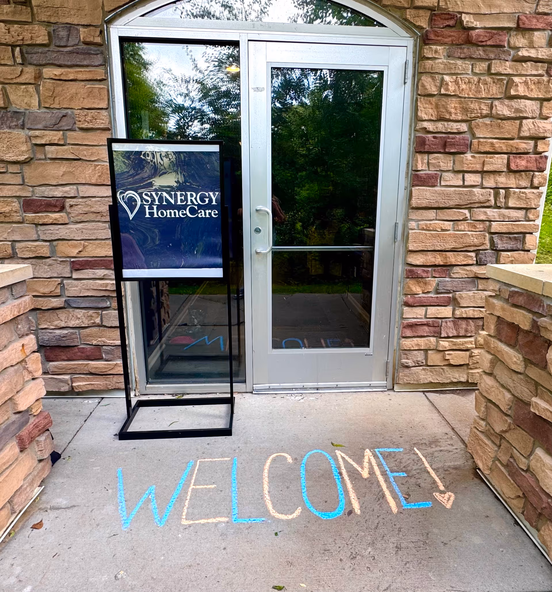 Glass front door of Synergy HomeCare entrance with colorful Welcome chalk on concrete.