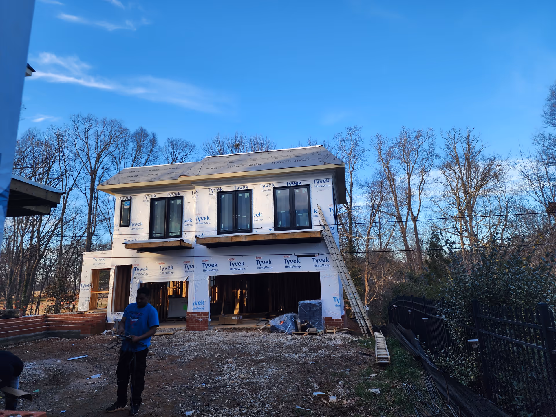 Two-story house under construction with Tyvek wrap, bare yard, and a worker in foreground.