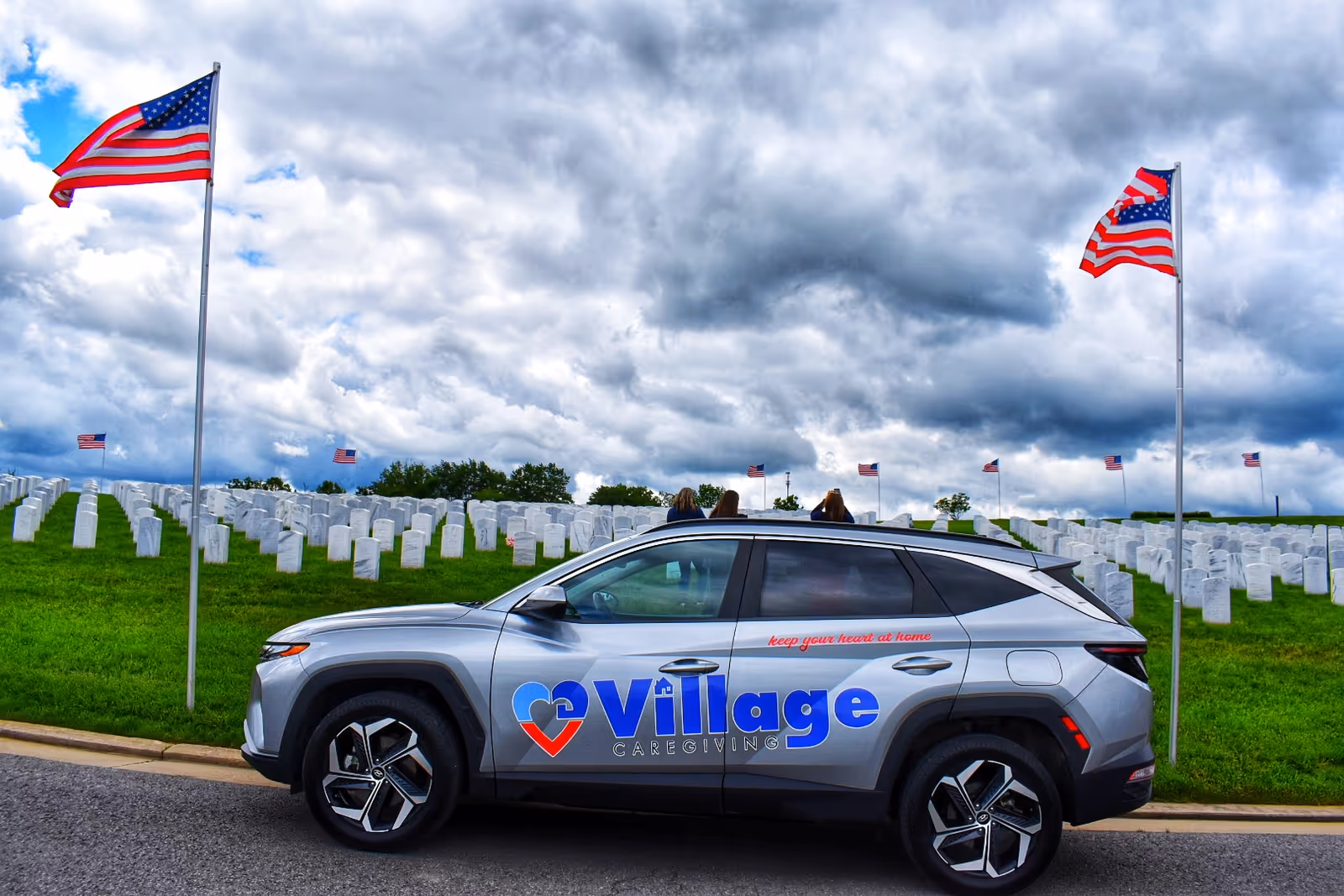 Silver Village Caregiving SUV parked by a cemetery with rows of white headstones and flags.