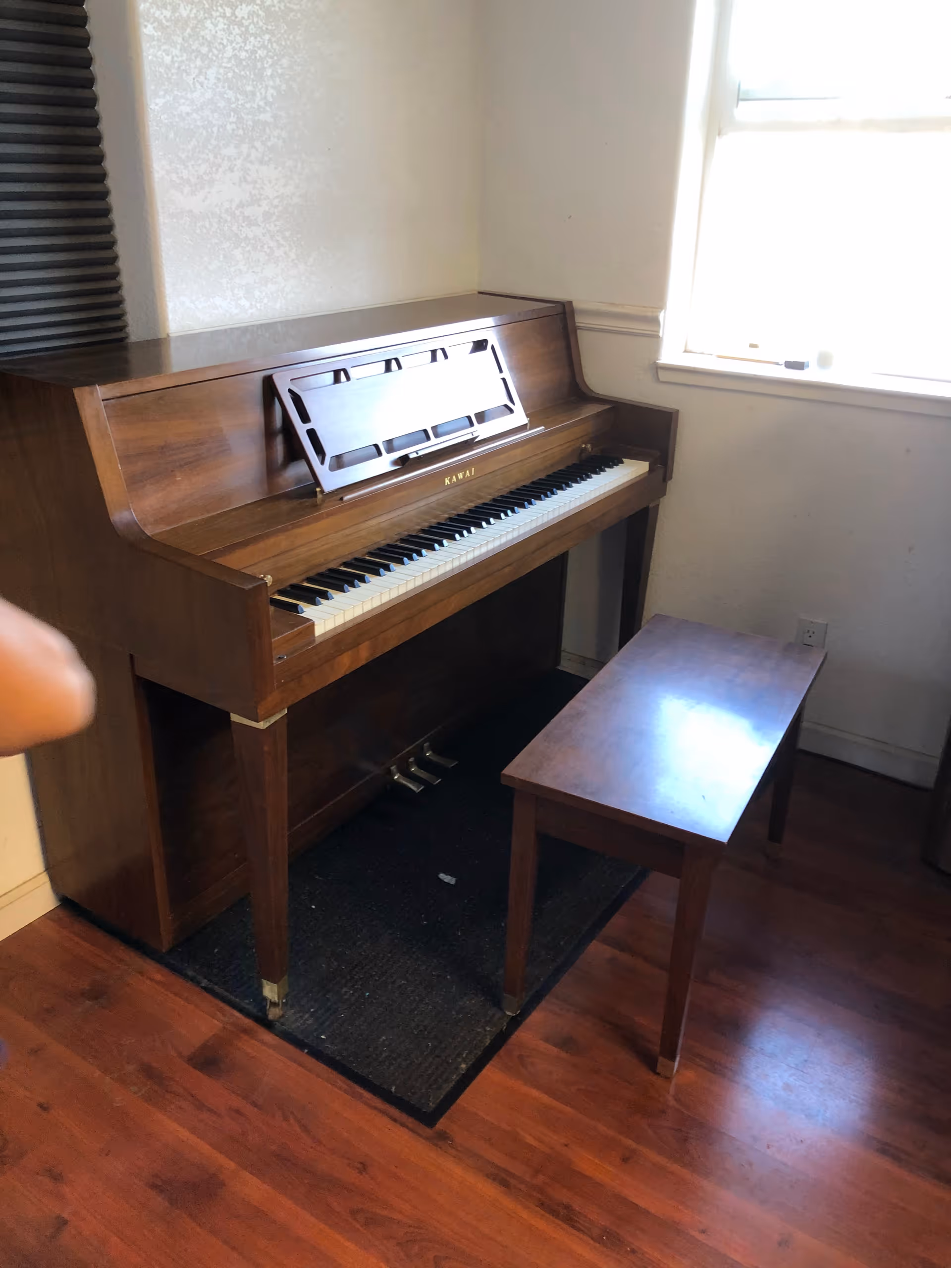 Wooden upright piano with bench sits in a small lit room near a window.