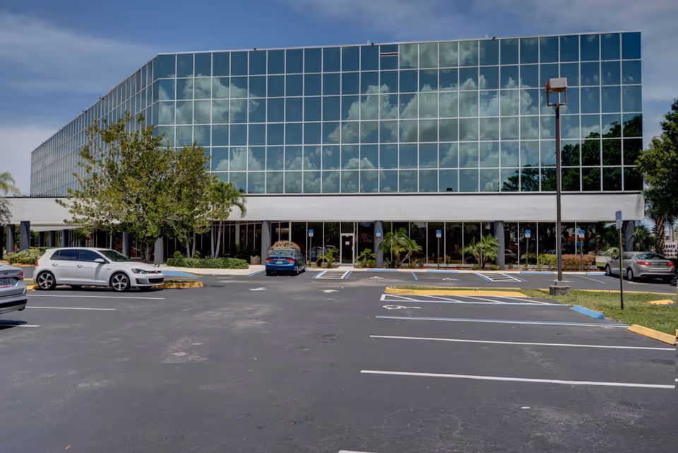 Modern glass-front building with a parking lot and trees in front