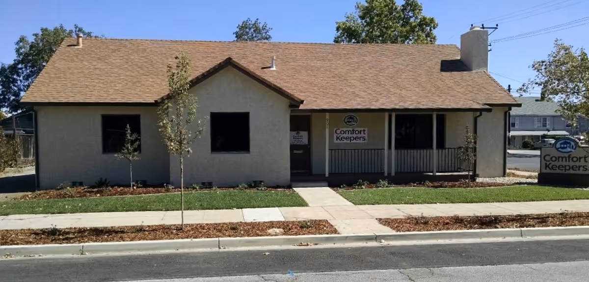 A single-story beige building with a brown roof and a Comfort Keepers sign beside a sidewalk.
