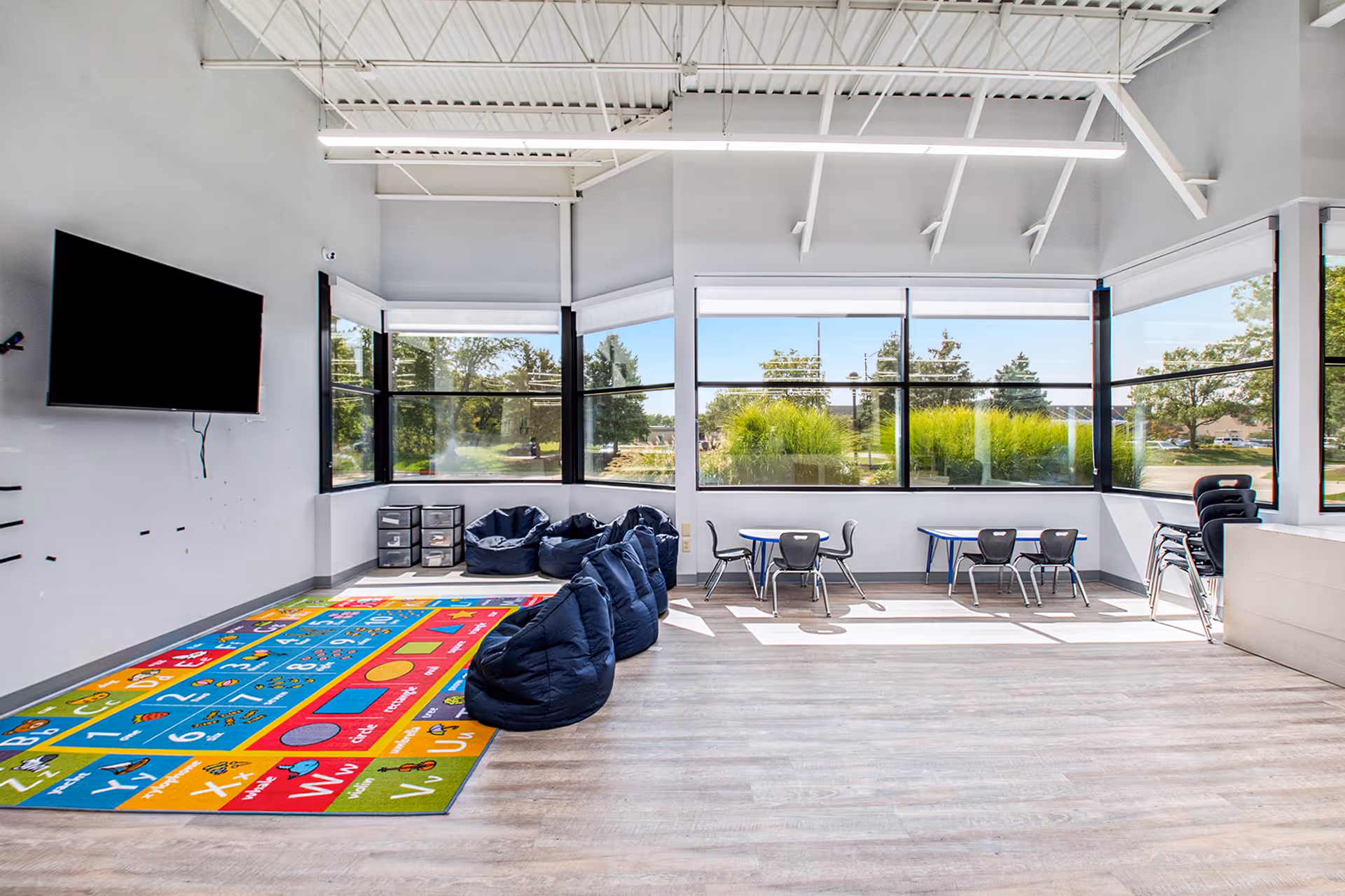 Bright activity room with colorful rug, bean bags, tables, chairs, and wall-mounted TV.