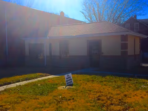 Small single-story building with a sloped roof, yard, and a sign in the grass.