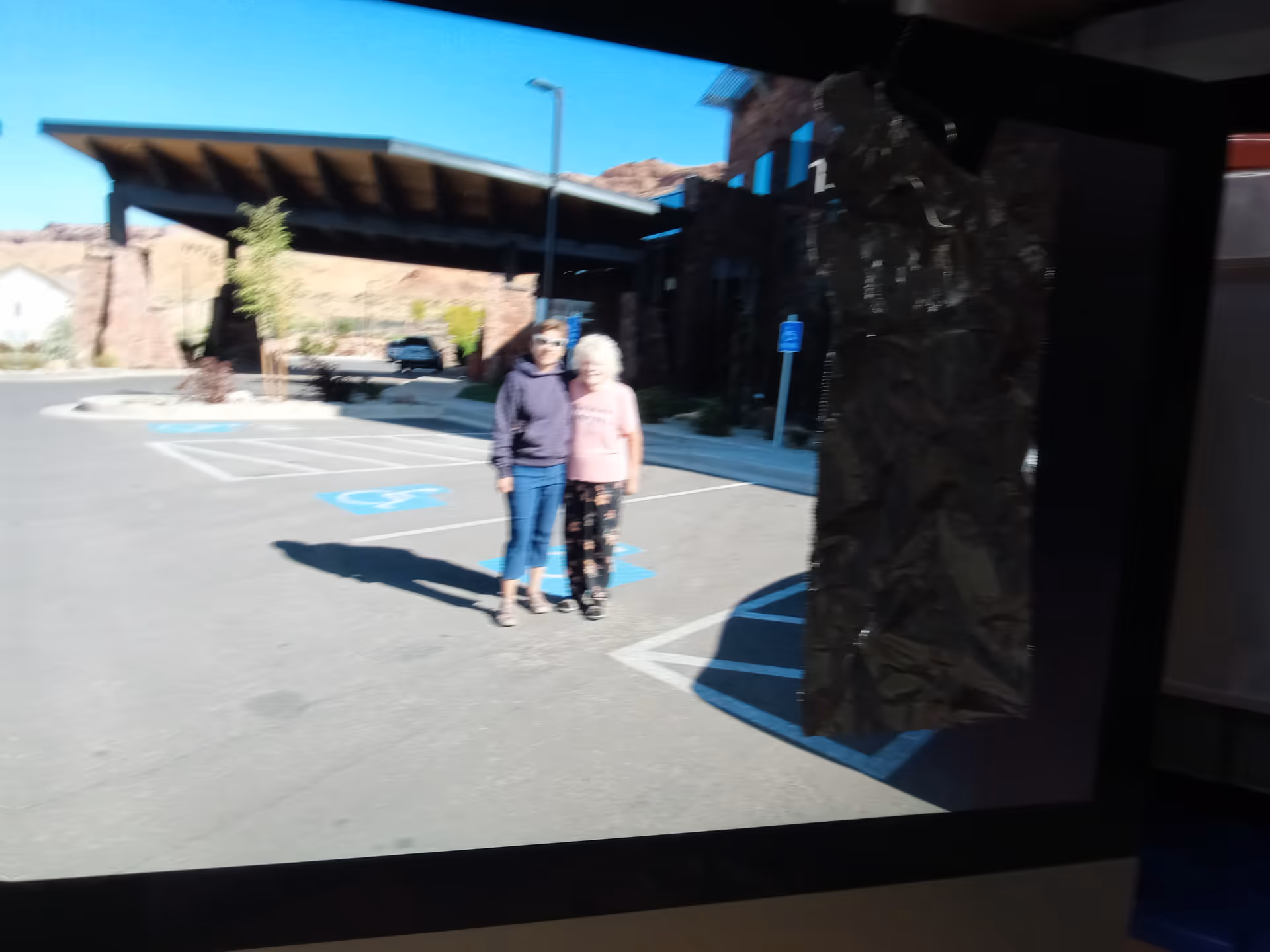 Two elderly women stand together in a parking lot near a building with a covered entrance.