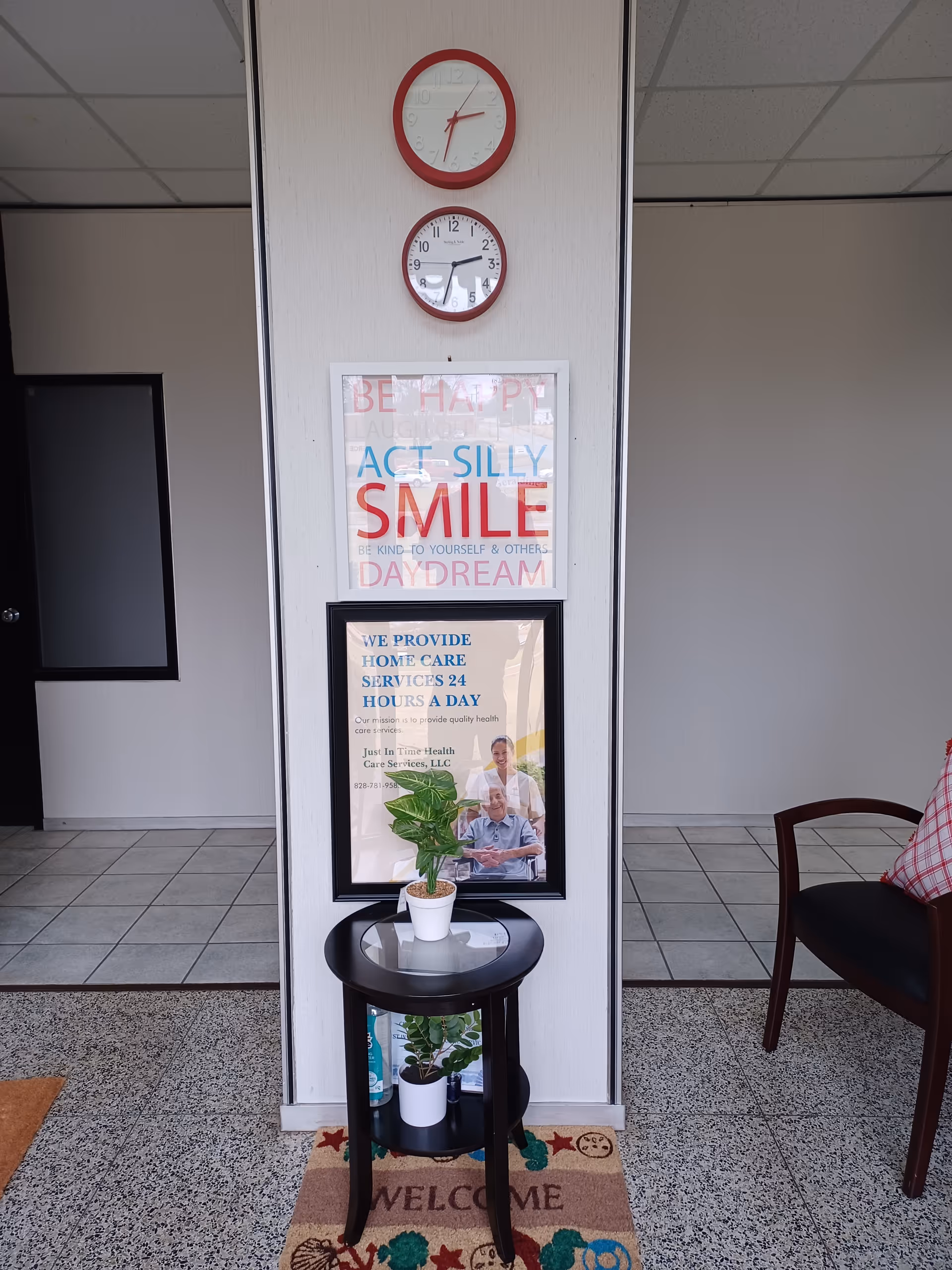 Interior lobby area with two wall clocks, framed signs, a table plant, and a welcome mat.