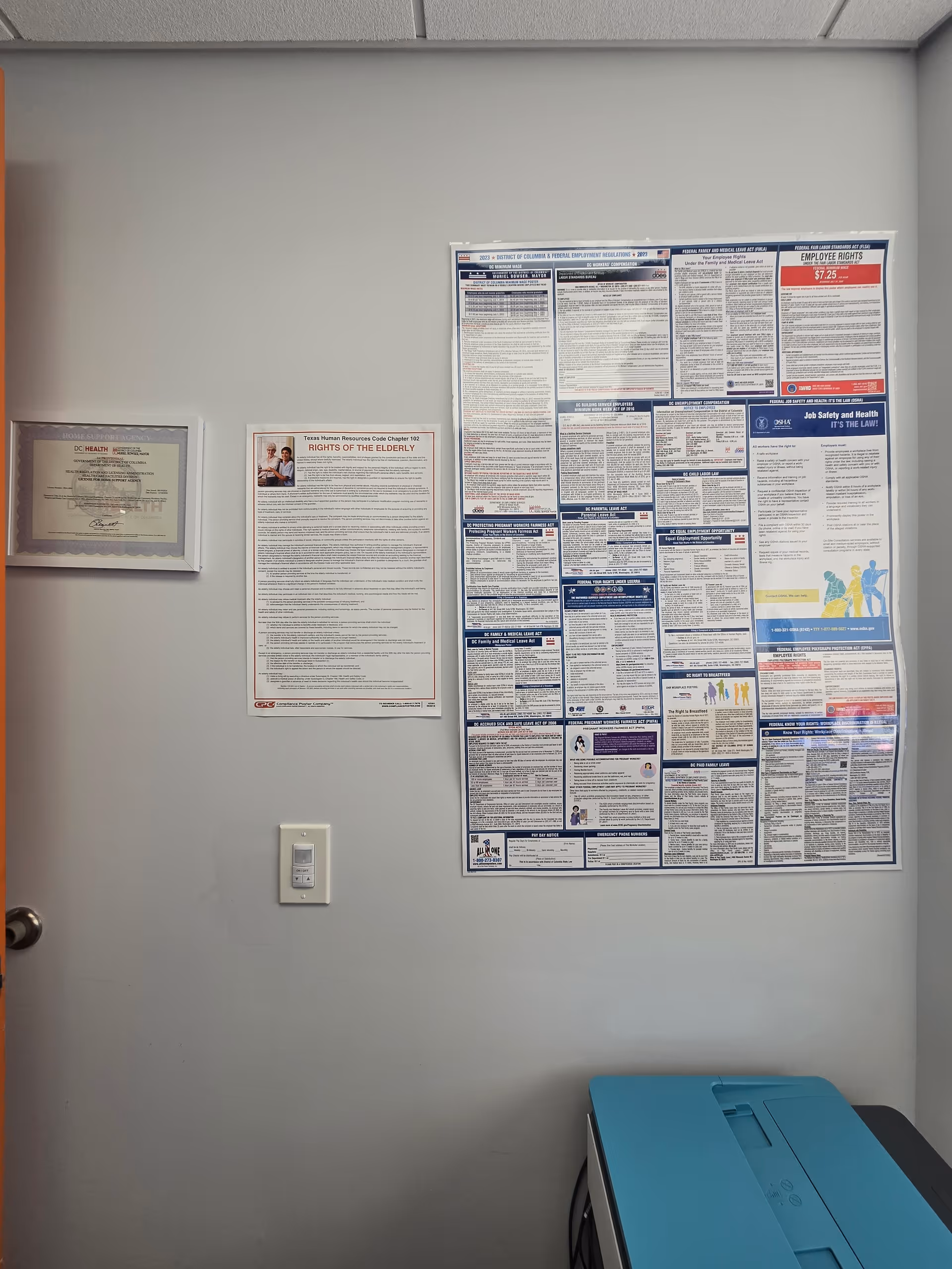 Gray wall with framed documents and a large bulletin board beside a teal printer.