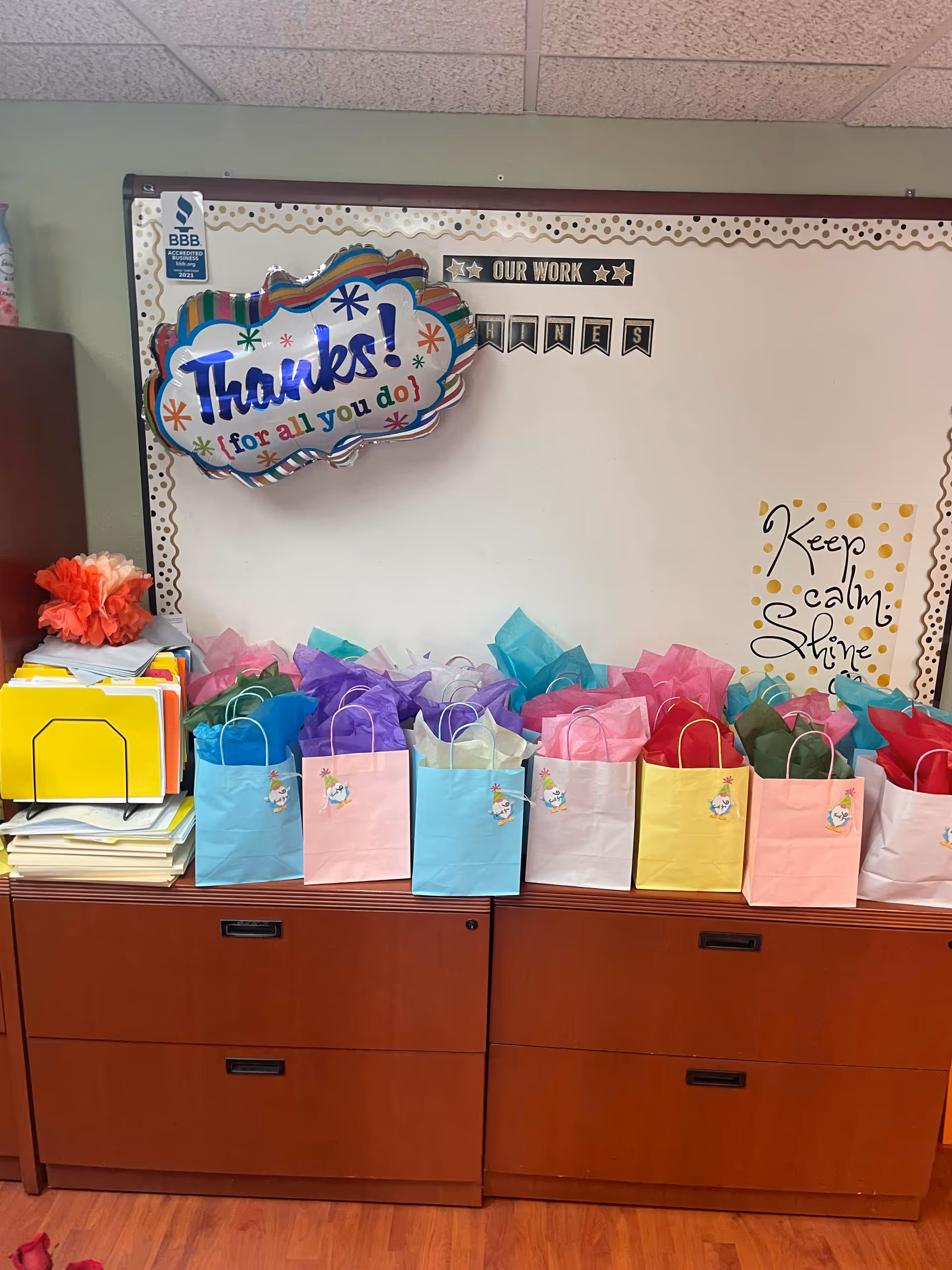 Colorful gift bags line a cabinet beneath a cheerful 'Thanks!' bulletin board in an indoor facility.
