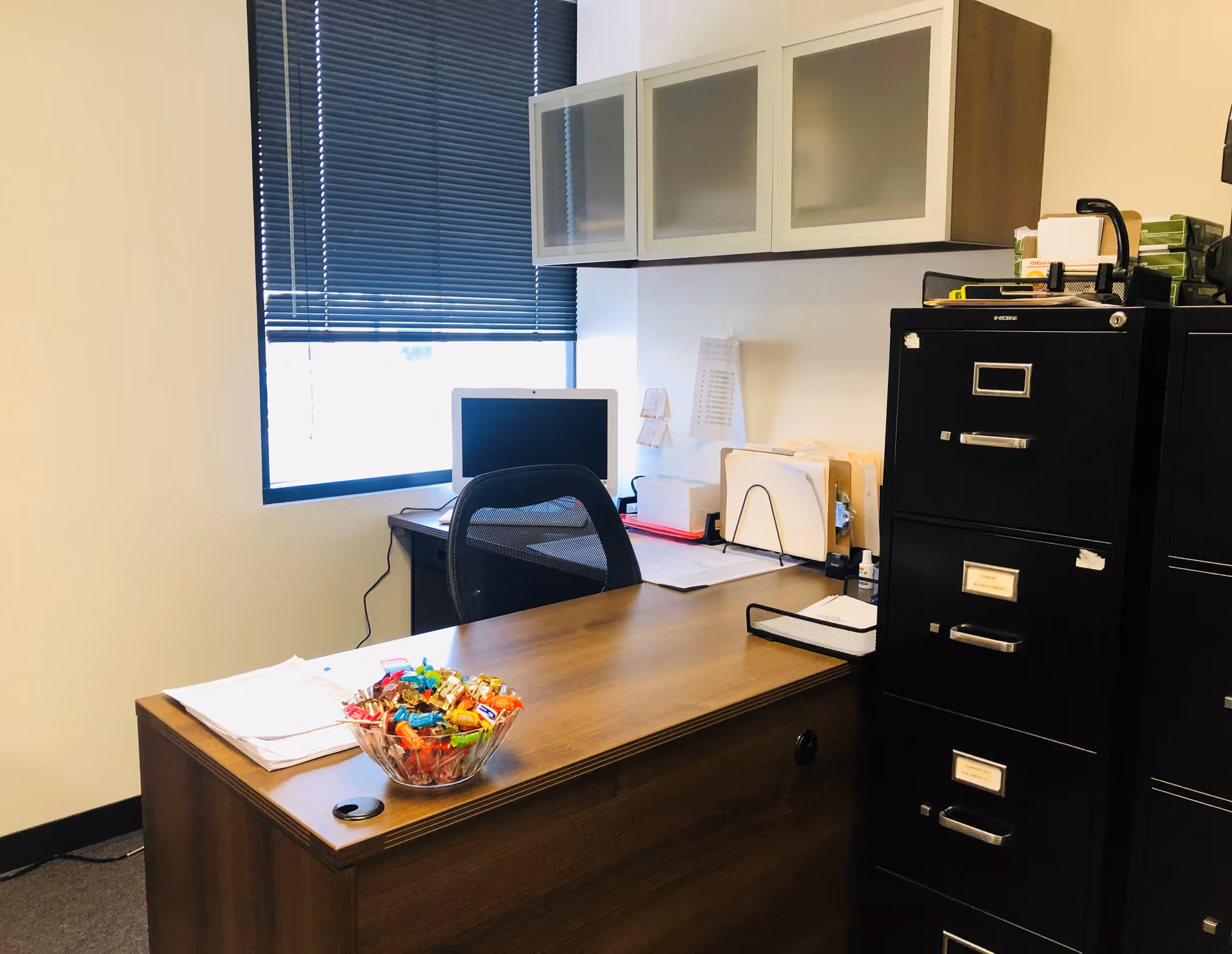 Office workspace with wooden desk, chair, computer, filing cabinets, and candy bowl under window blinds.