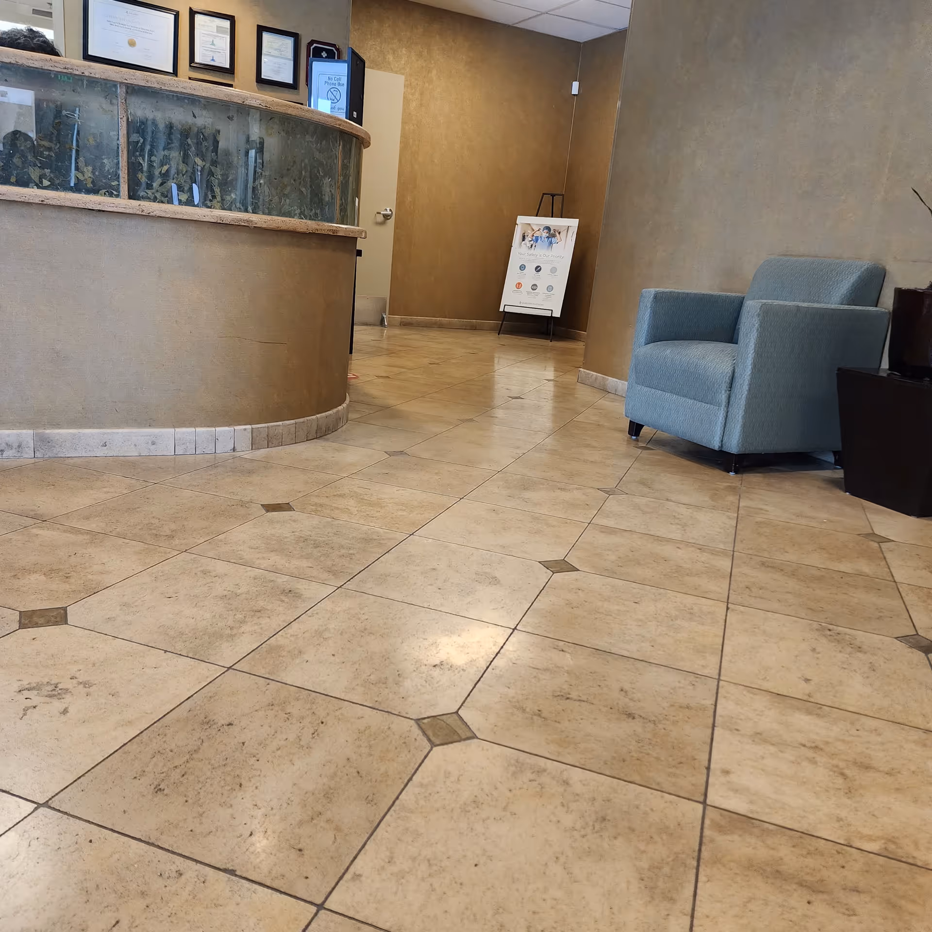 Interior of a lobby with curved reception desk, framed certificates, tiled floor, and blue armchair.