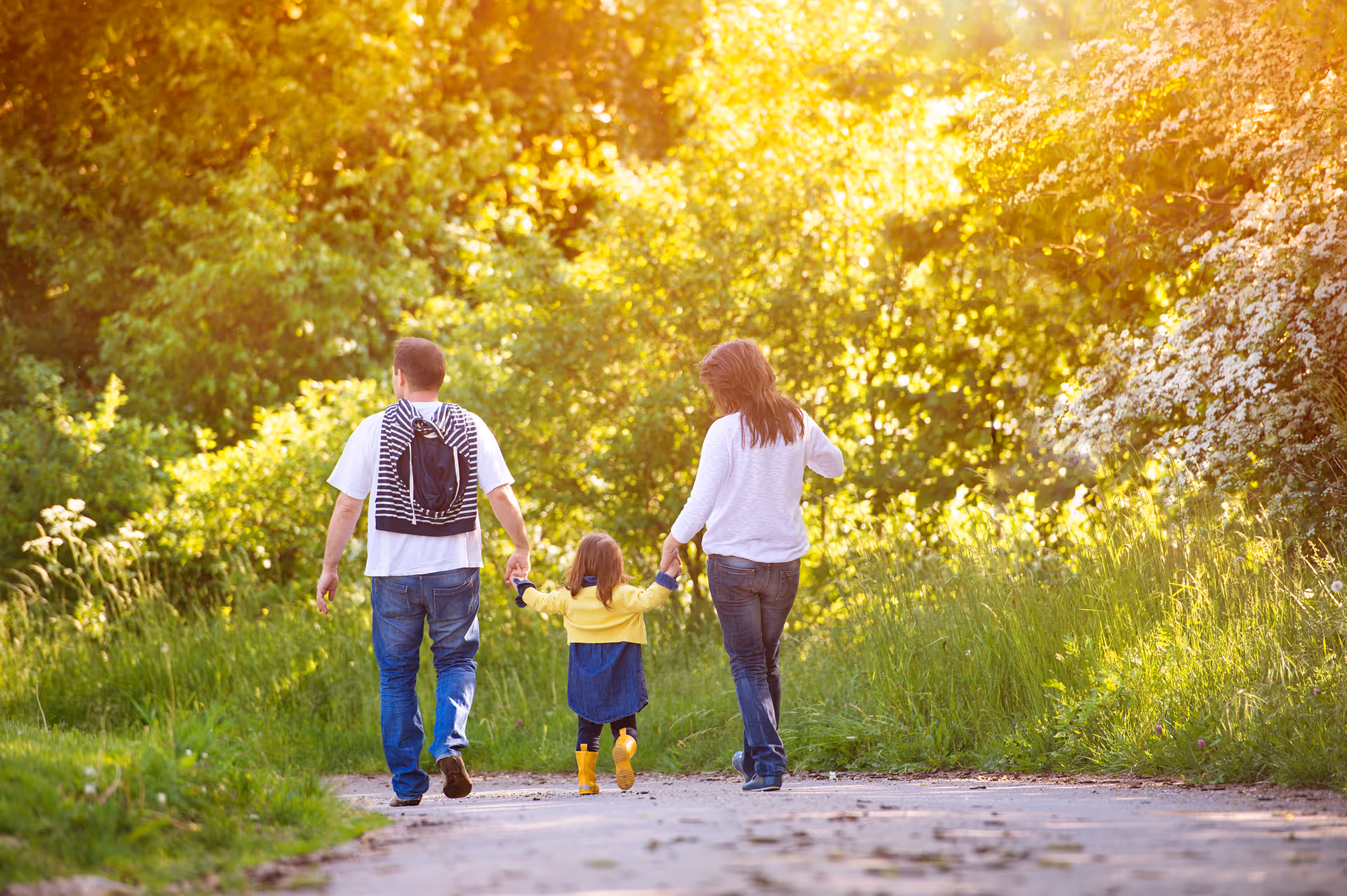 Family of two adults and a child walk hand-in-hand along a sunlit park path.