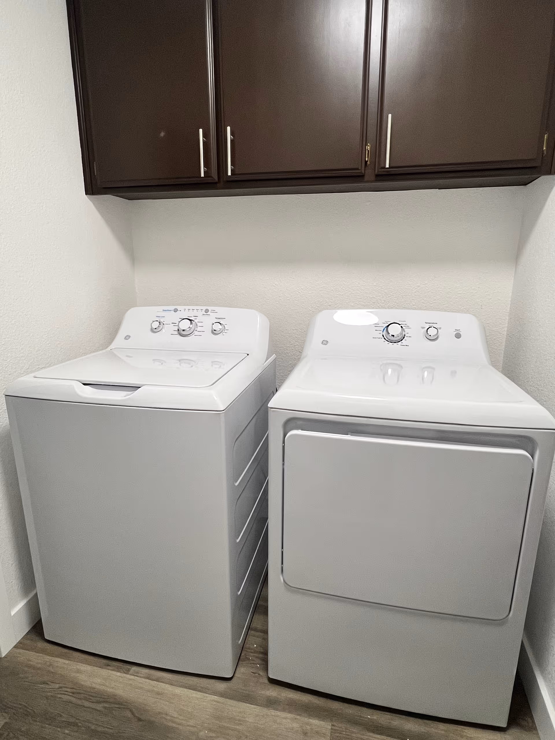 White washing machine and dryer sit side by side under dark cabinetry in a laundry room.