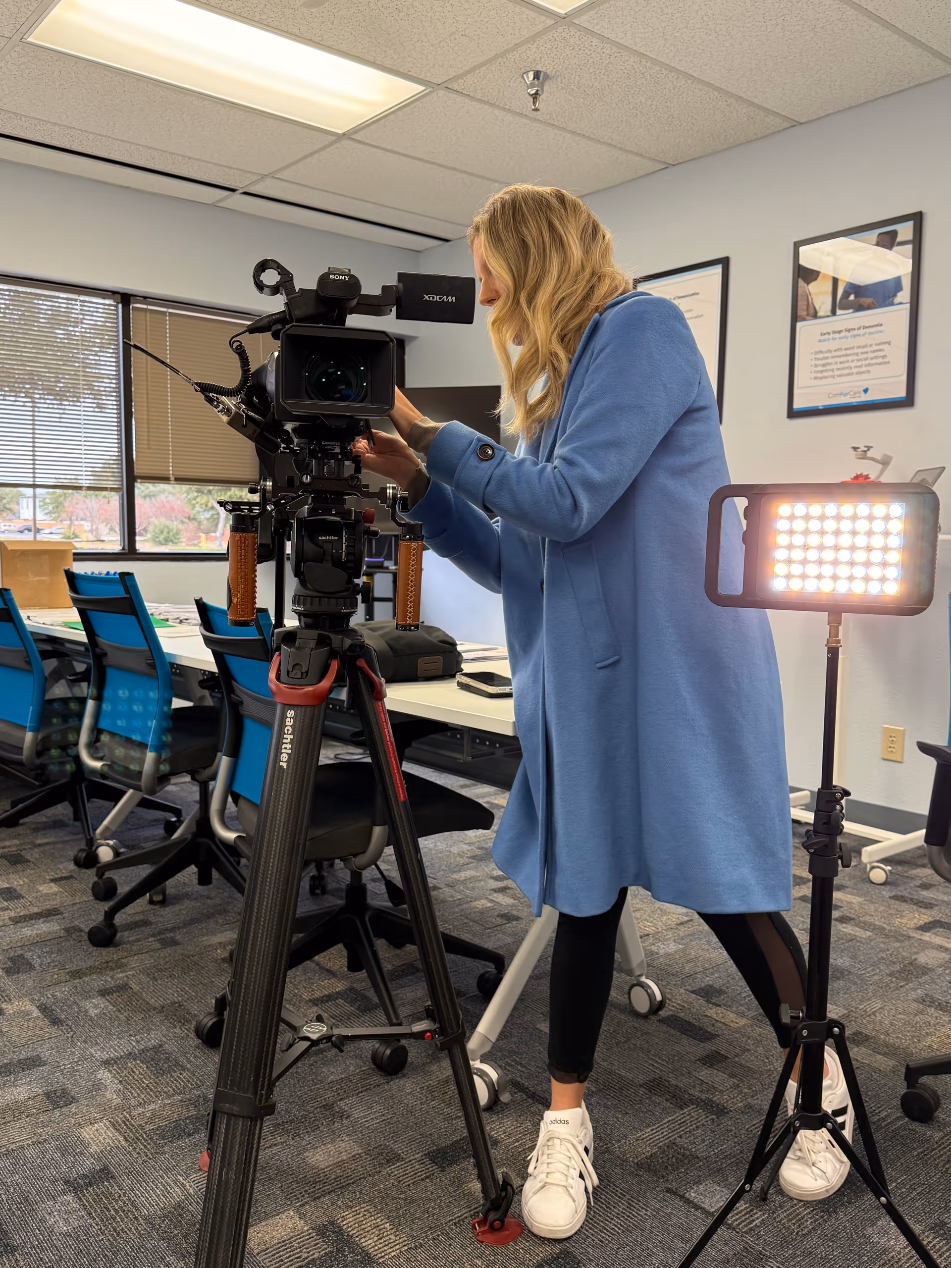 A woman operates a professional video camera on a tripod in a bright conference room.