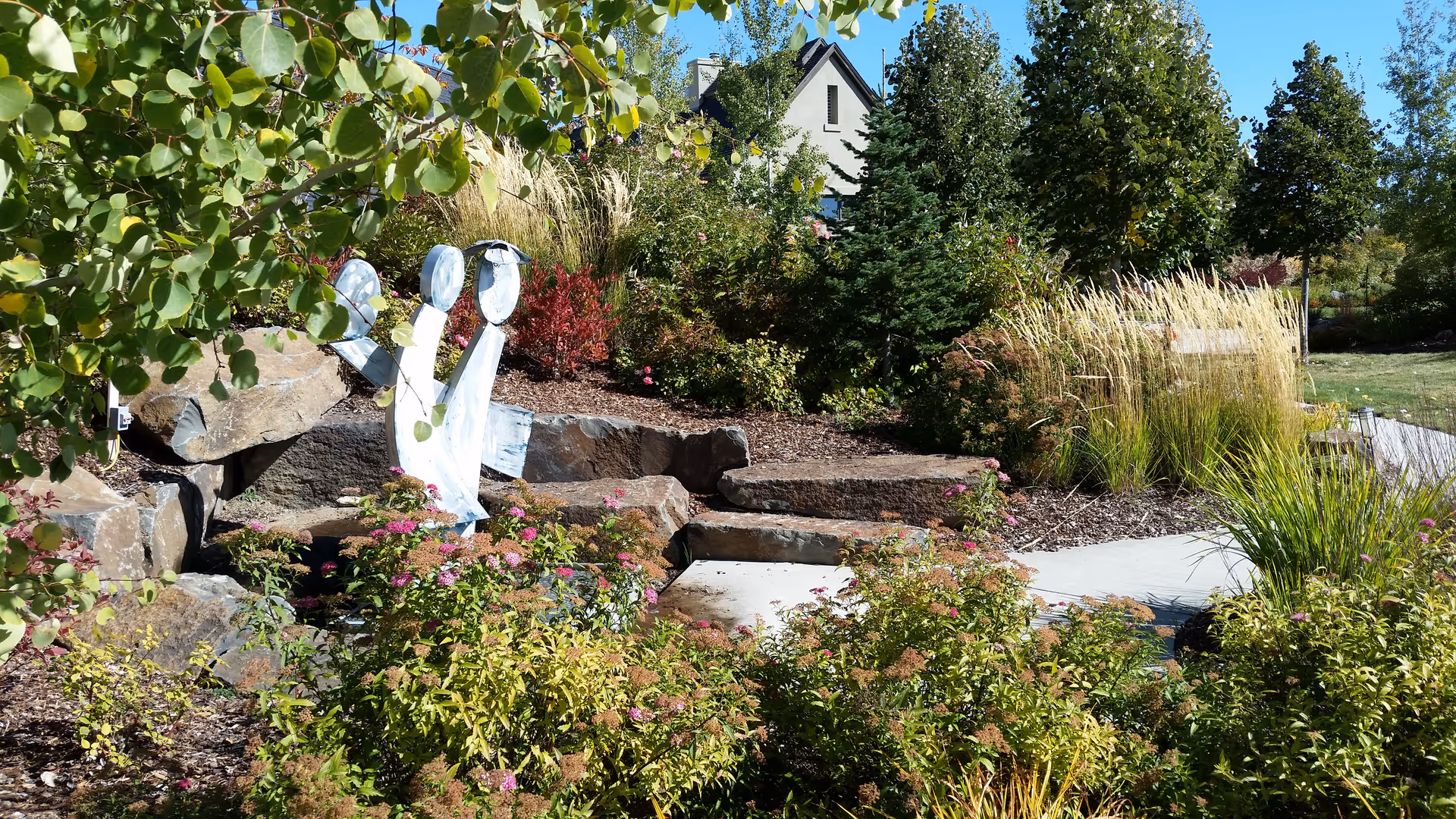 A landscaped outdoor garden with rocks, shrubs, ornamental grasses, and a white sculpture.