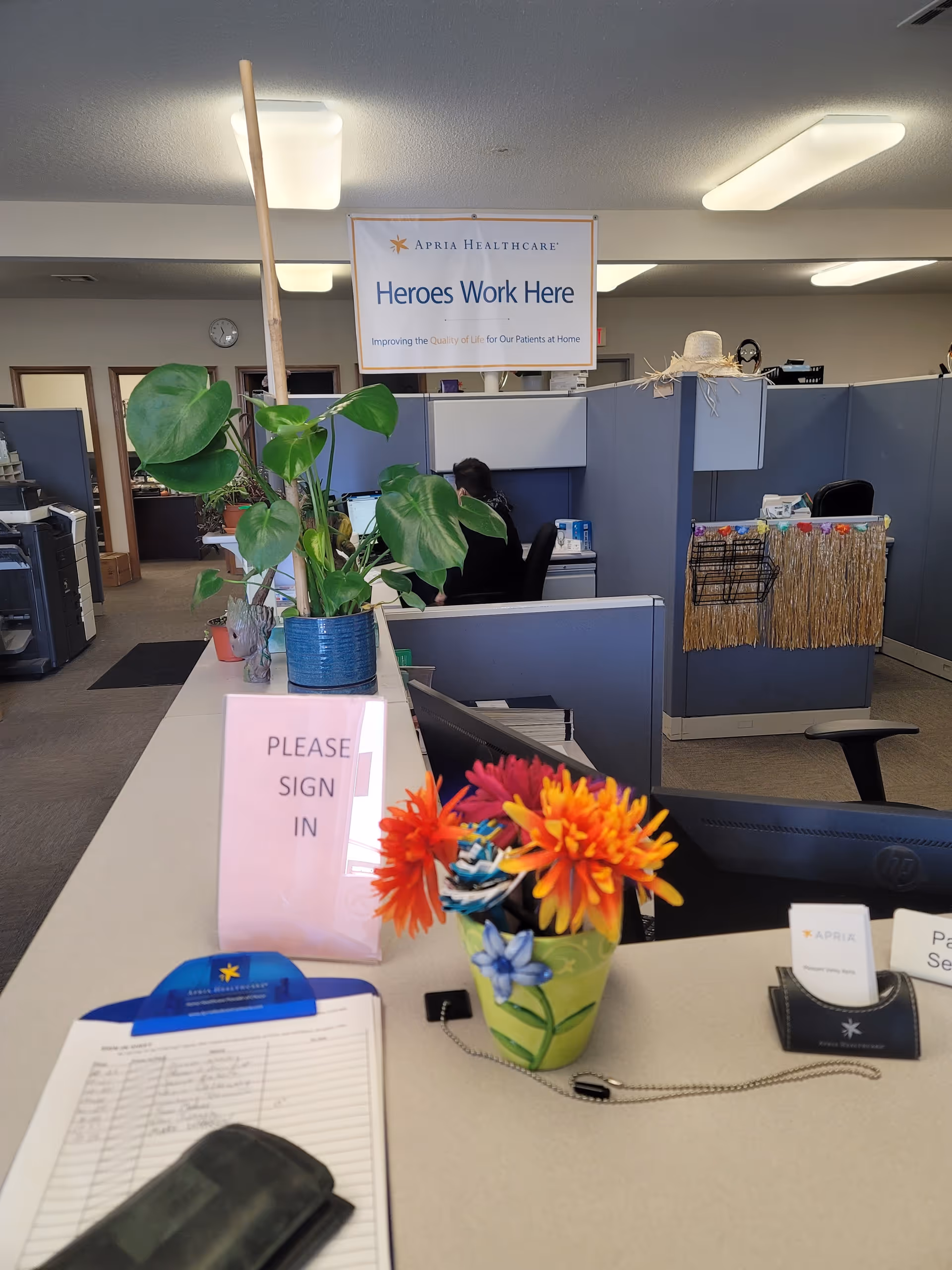 Reception desk at Apria Healthcare with sign-in, plants, and cubicle partitions inside.