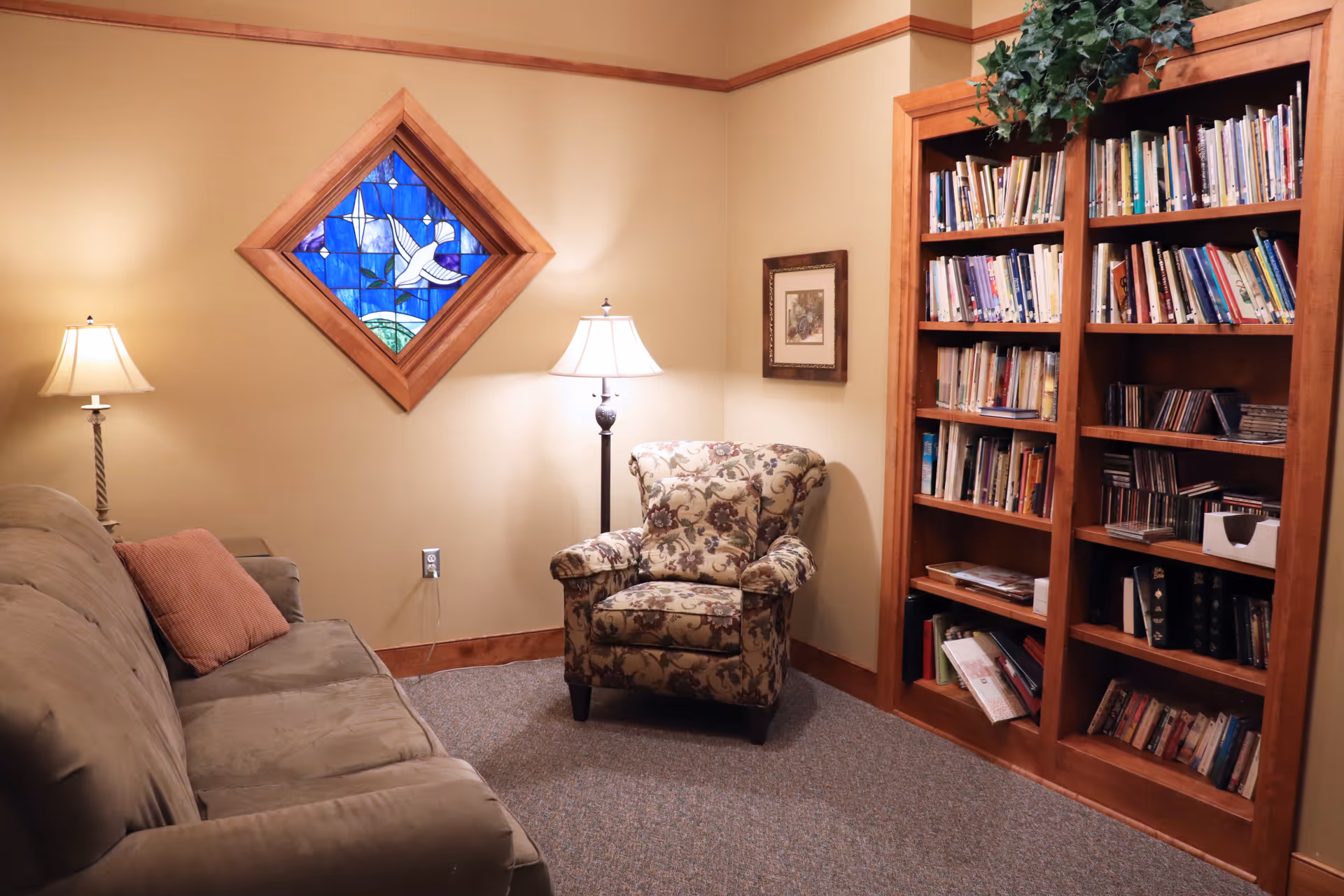 Cozy lounge area with beige walls, armchair, sofa, lamps, stained glass, and bookshelf.