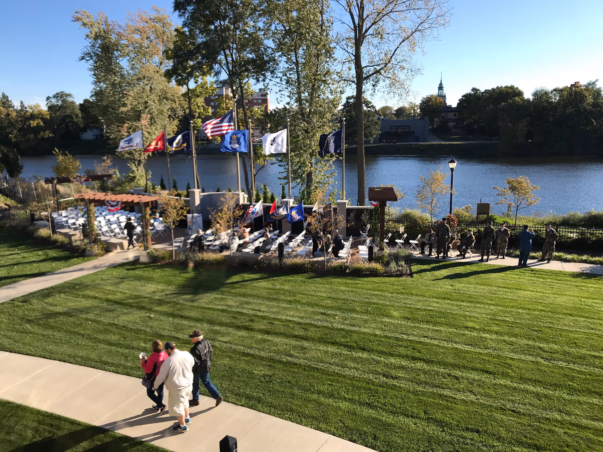 Outdoor riverside ceremony area with flags, rows of chairs, trees, and people walking.