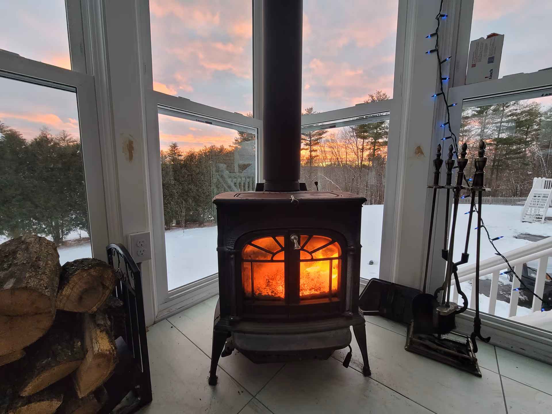 A wood-burning stove glows warmly in a bright sunroom with snowy outdoors.