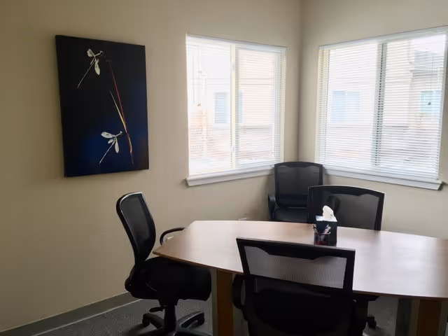 Small meeting room with a wooden table, black chairs, windows, and wall art.