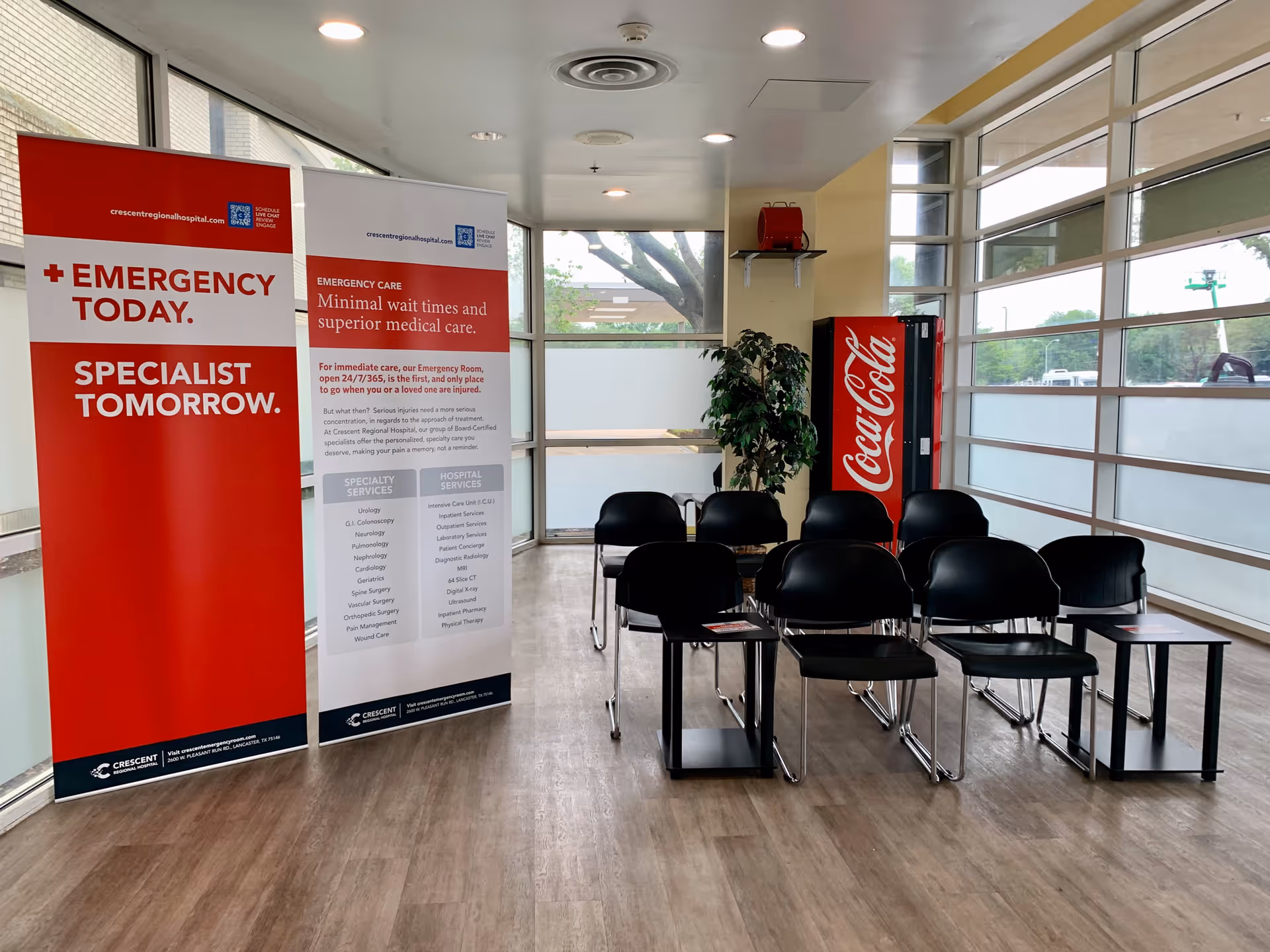 Interior hospital lobby with black chairs, red banners, a Coca-Cola vending machine, and windows.