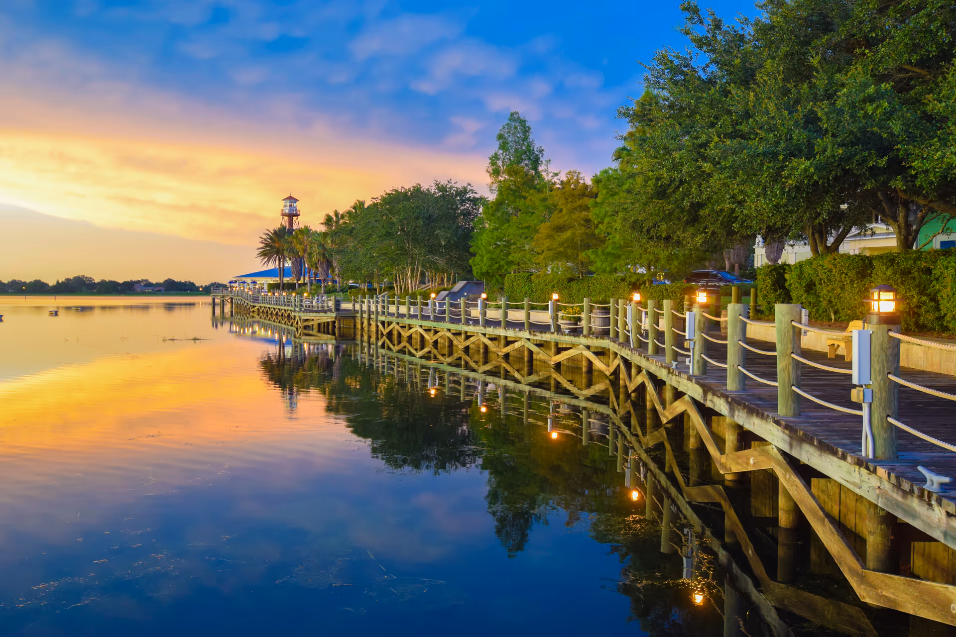 Sunset-lit wooden boardwalk along a calm lake with trees and lampposts.