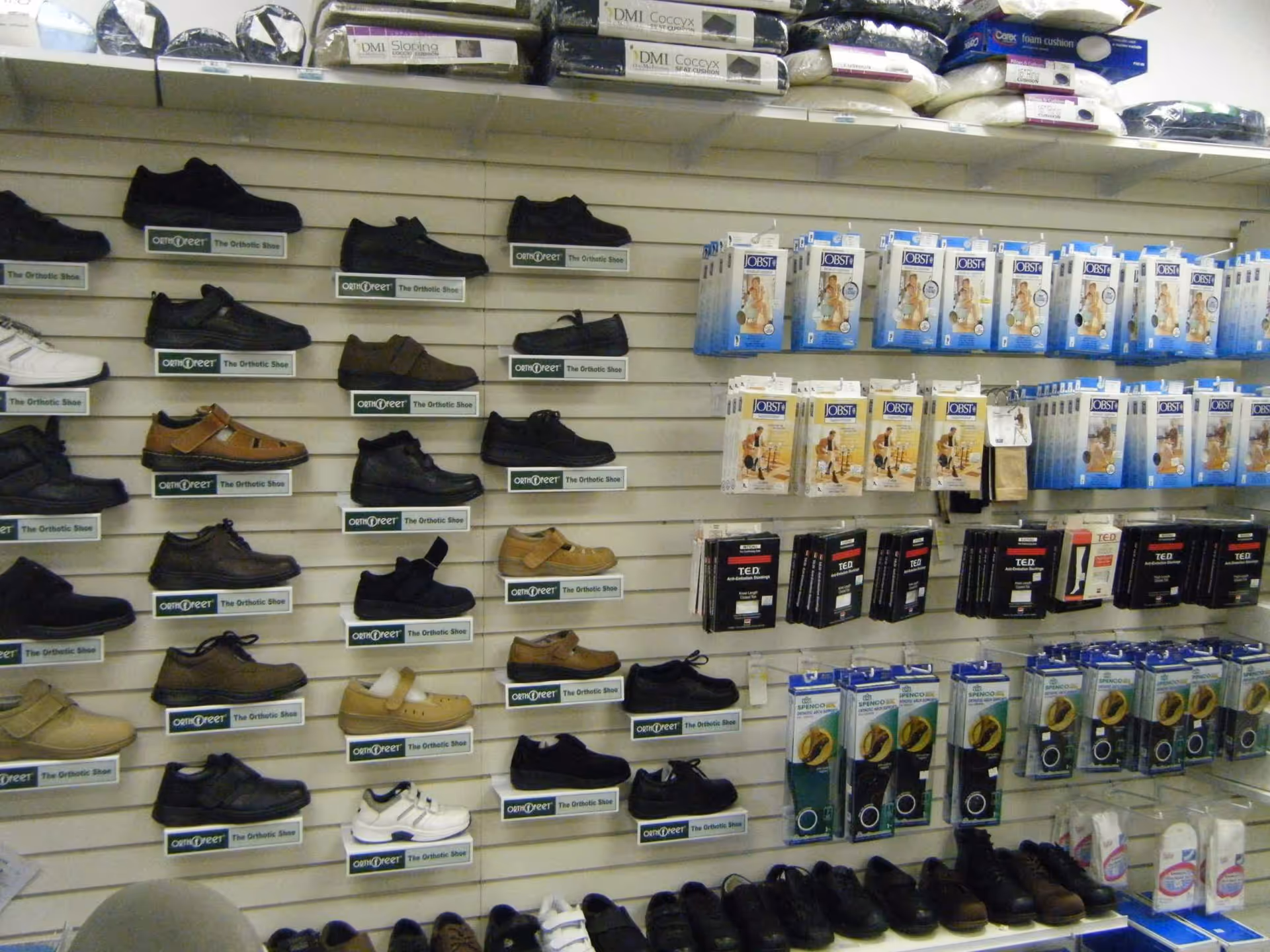 Wall shelves display numerous orthopedic shoes and footwear accessories in a retail shop.