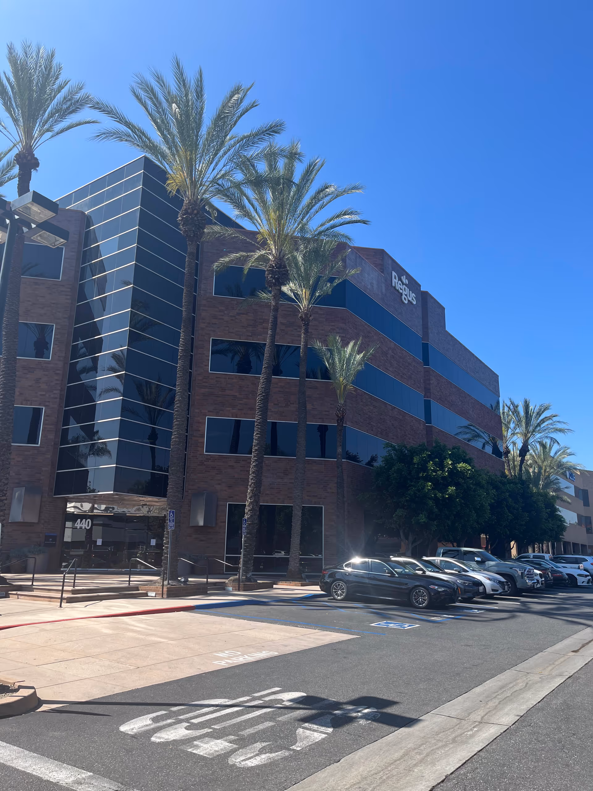 Modern brick building with glass windows, palm trees, and parked cars along the street.