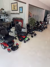 Row of mobility scooters and power chairs lined up inside a showroom with glass partitions.