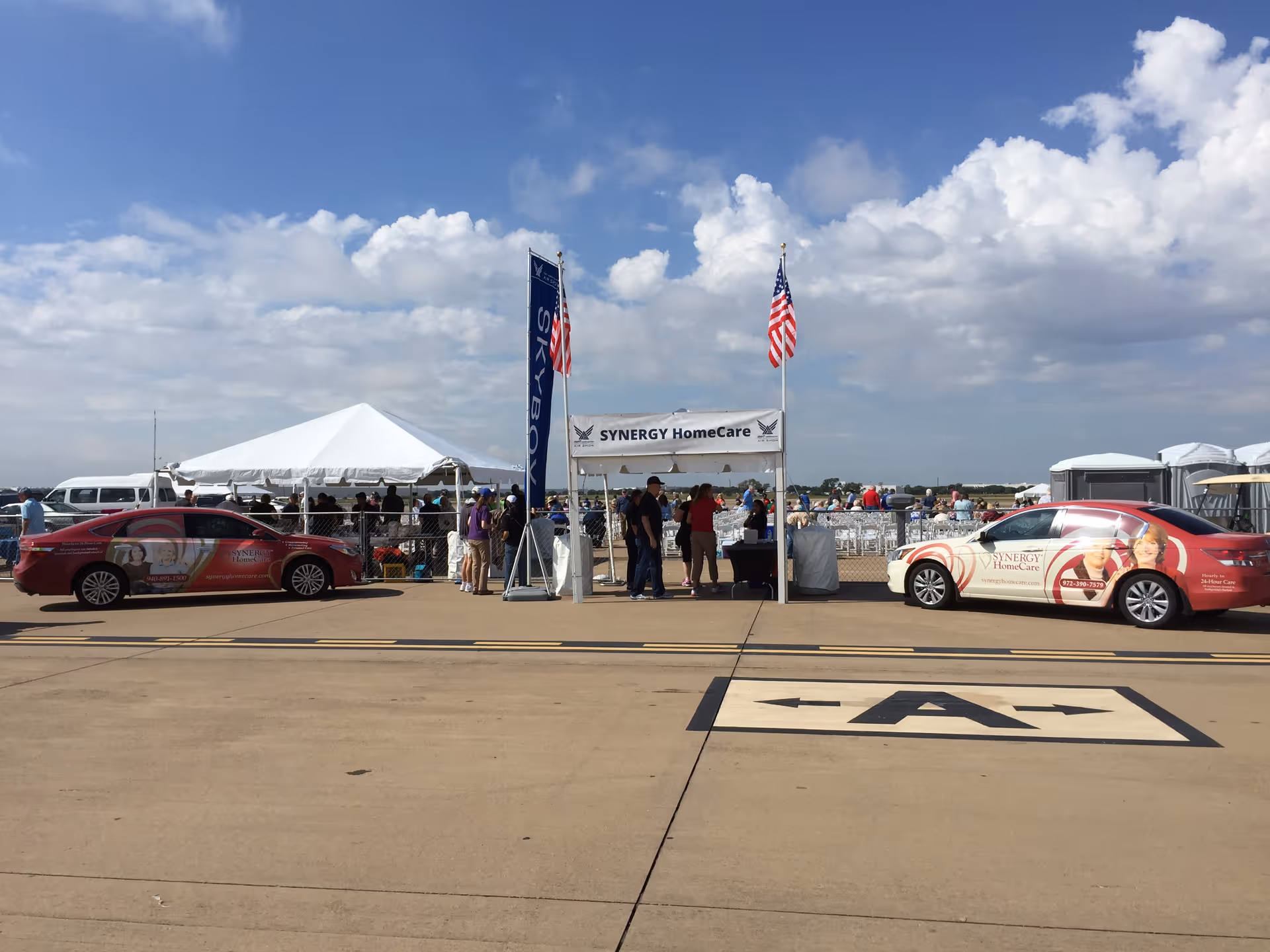 Outdoor promotional event for SYNERGY HomeCare with tents, banners, and branded cars on tarmac