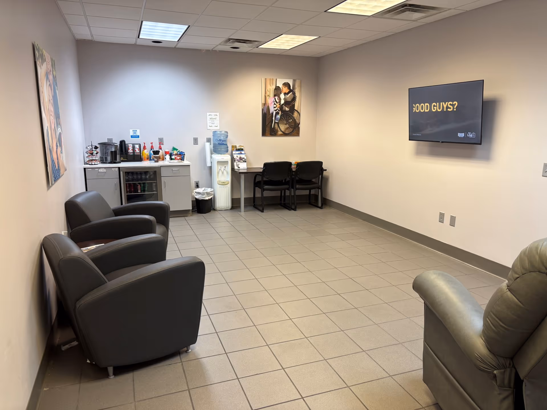 A spacious waiting area with gray chairs, a wall-mounted TV, and refreshments.