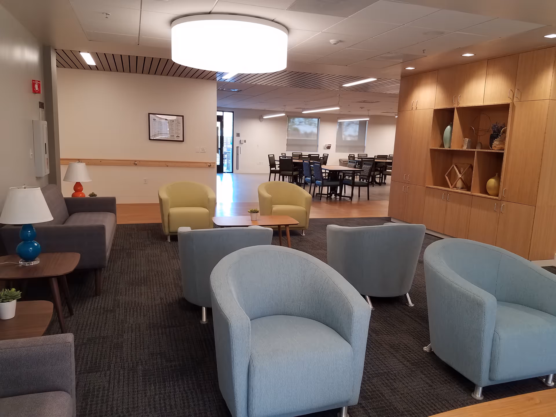 Cozy senior living lounge with light blue chairs, sofas, tables, and wooden cabinetry.