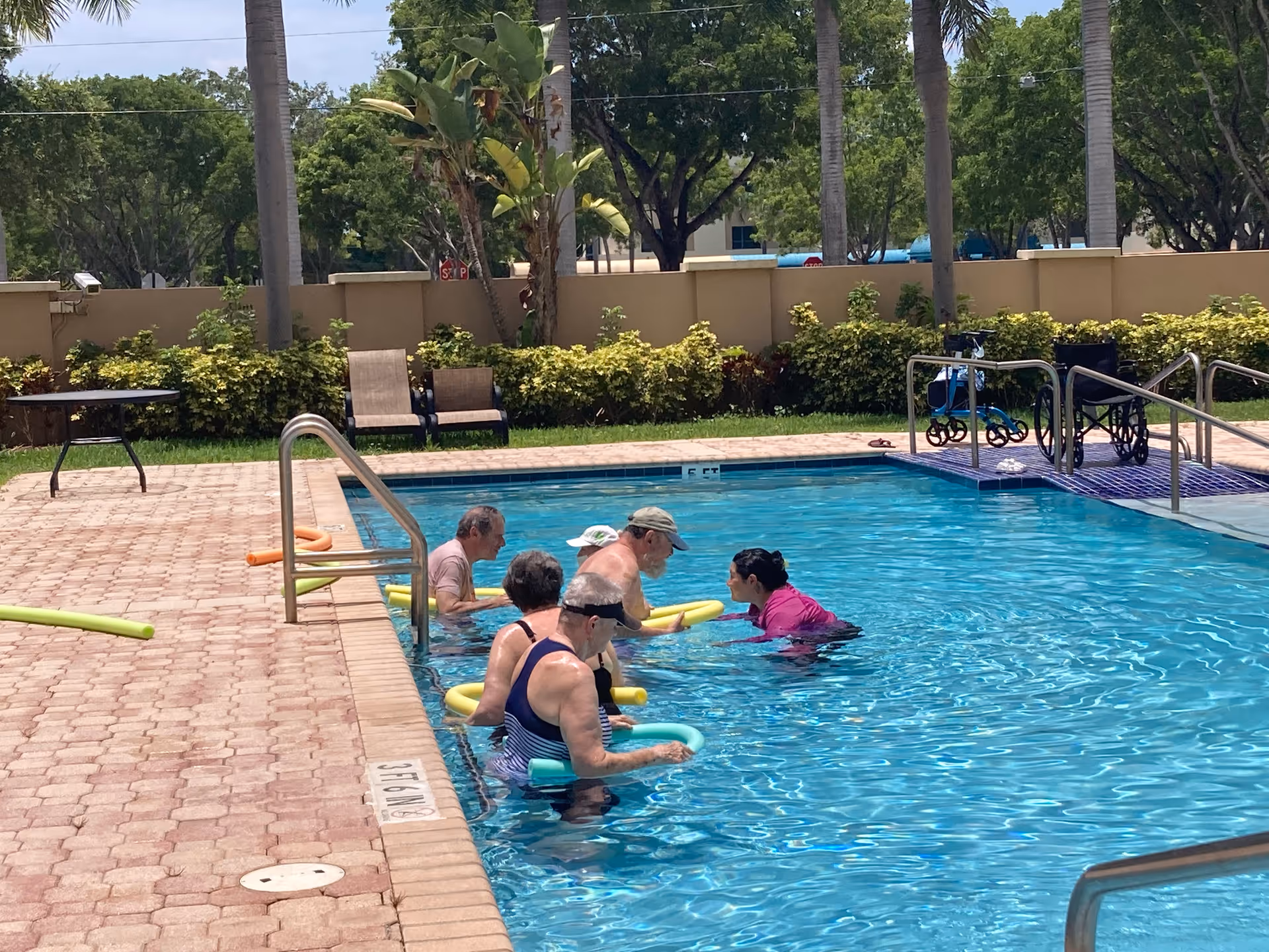 Senior residents swim with flotation noodles in an outdoor pool area.