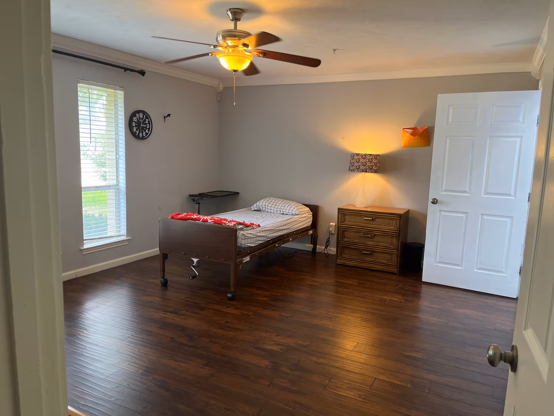 A sparsely furnished bedroom with a bed, dresser, lamp, and ceiling fan.