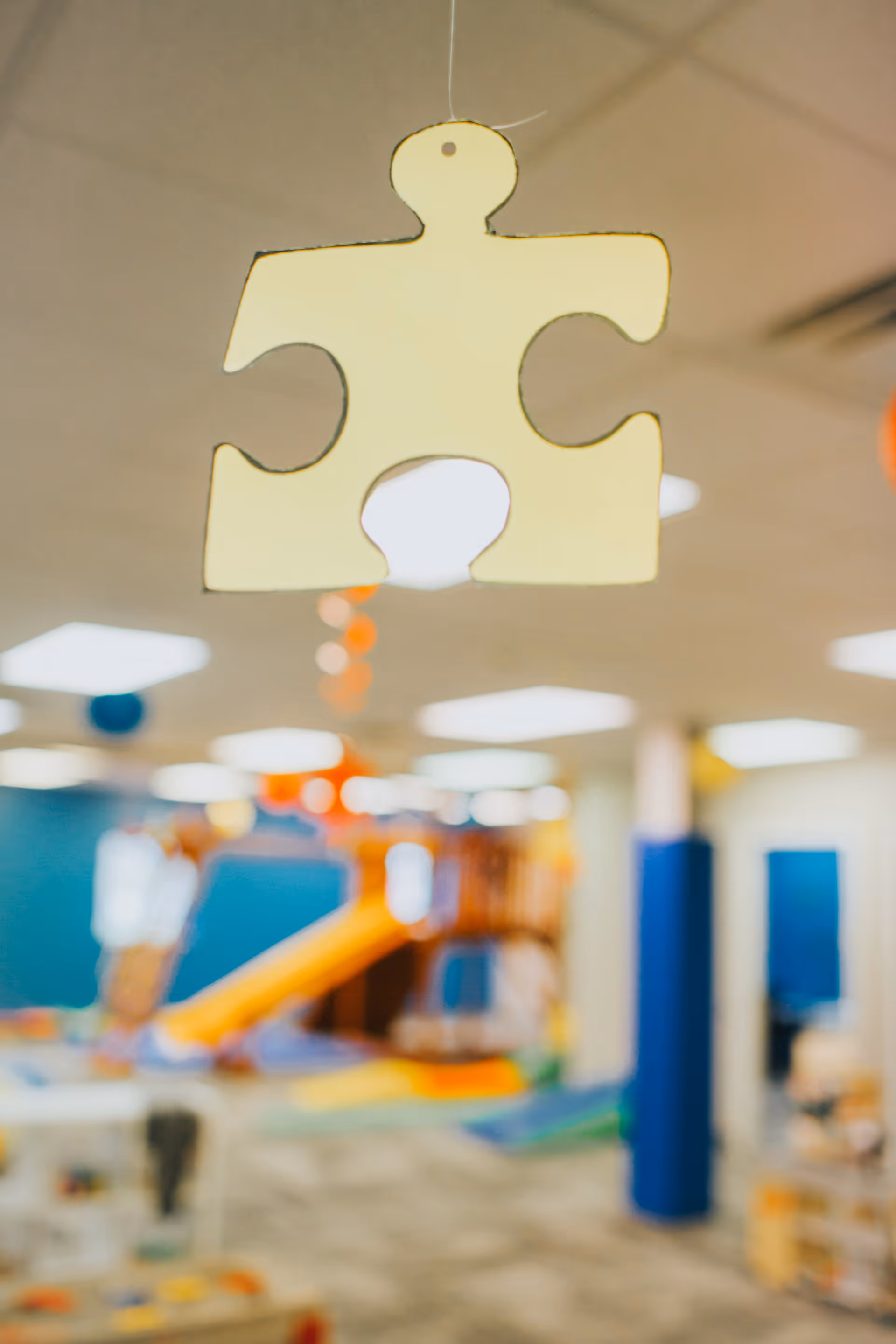 Hanging yellow puzzle-piece decoration in foreground with a colorful indoor play area blurred behind.