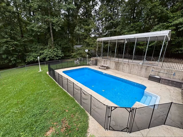 Outdoor pool area with a fenced lap pool, surrounding grass, trees, and a covered seating structure.