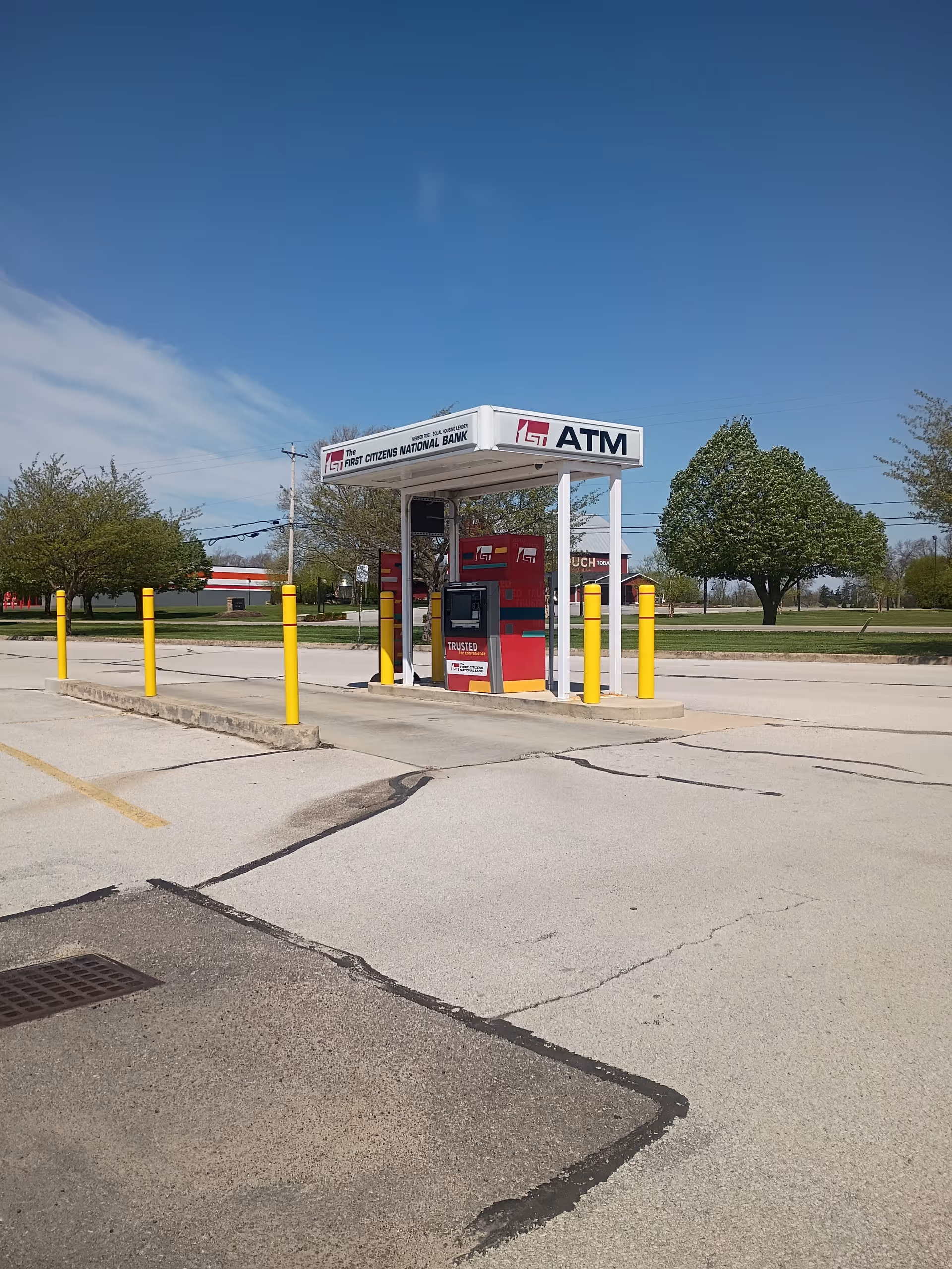 Outdoor parking lot with a white ATM kiosk surrounded by yellow bollards and trees