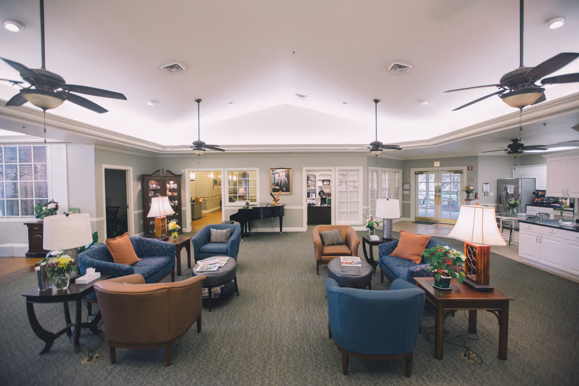 Spacious senior living lounge with sofas, tables, lamps, and a kitchen visible in background.