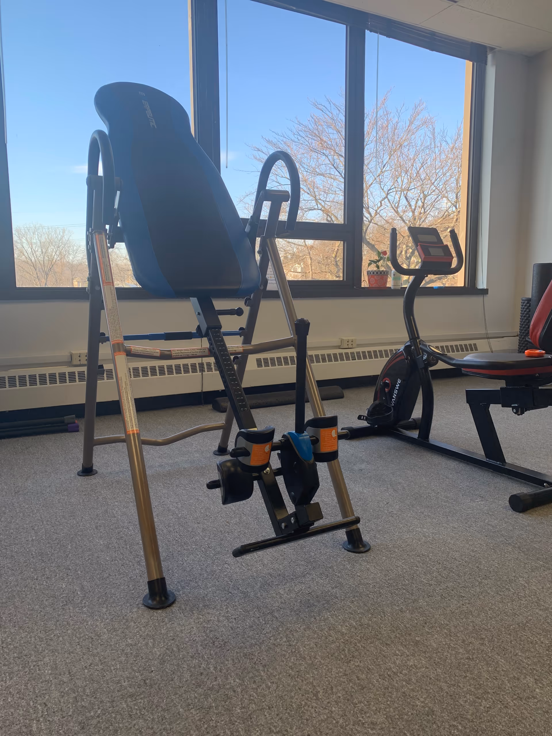 Interior fitness room with leg extension machine and stationary bike by large windows.