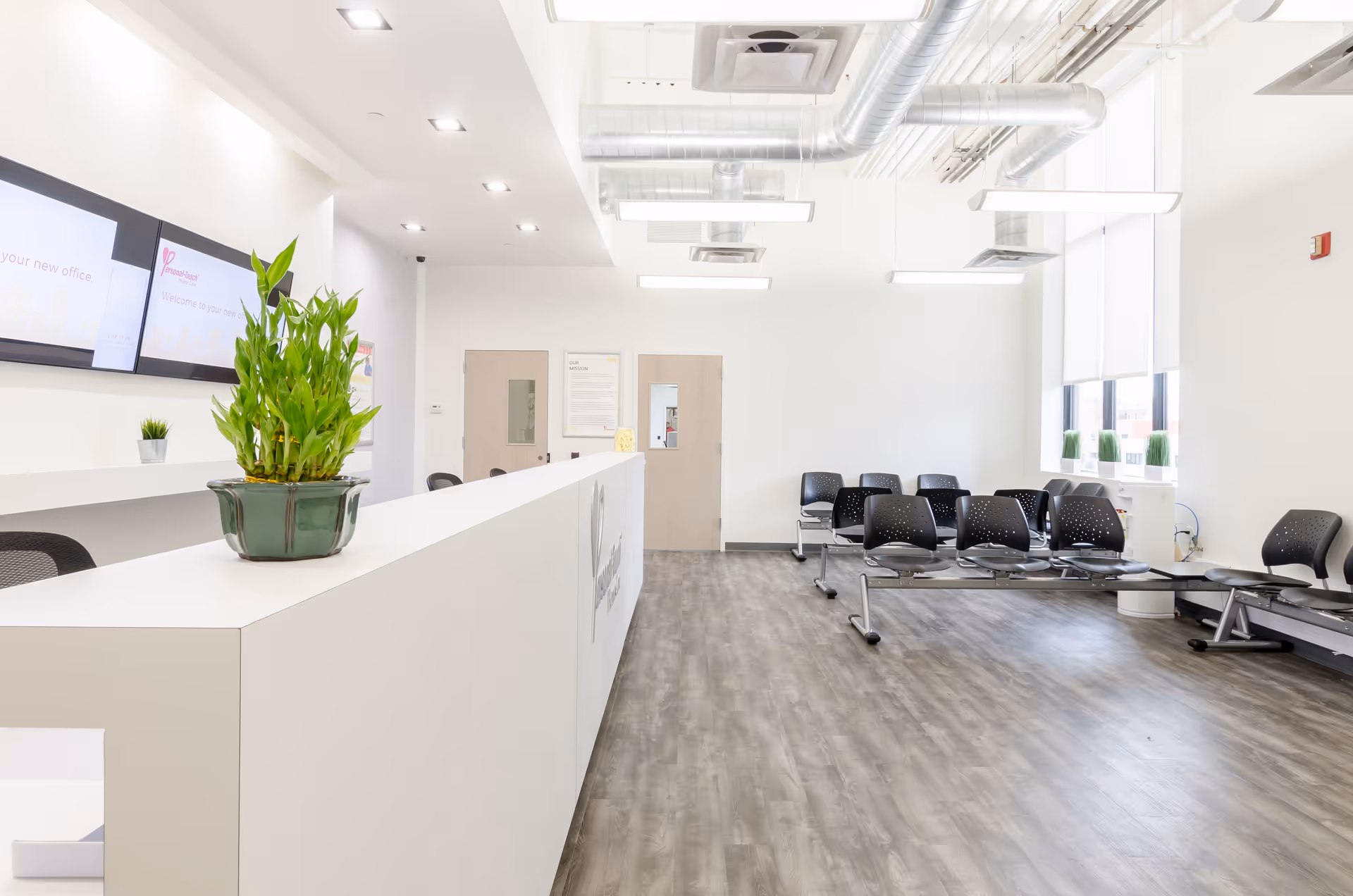 Bright reception area with a long white counter, green plant, and row of black chairs.