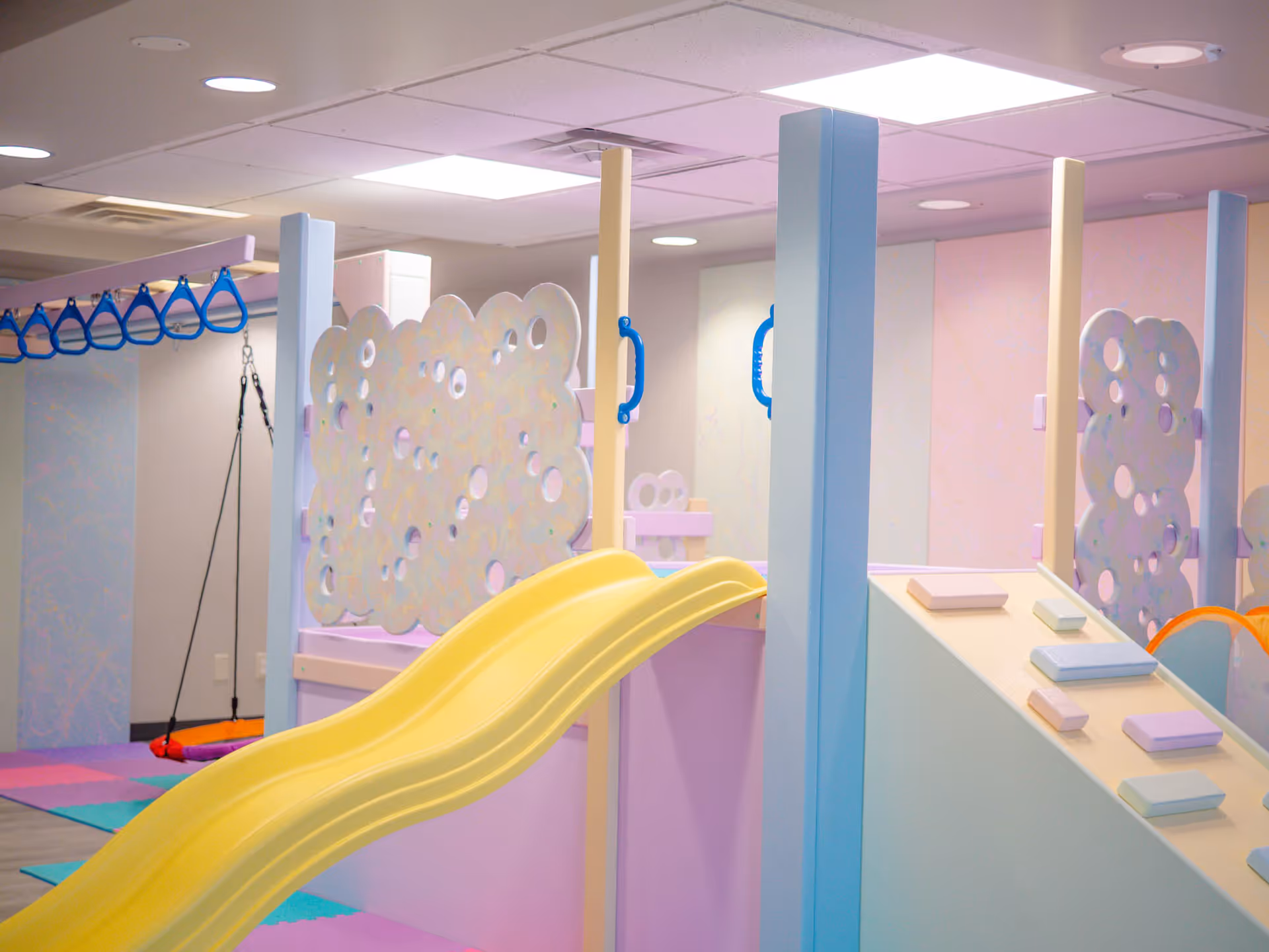 Bright pastel indoor play area with a yellow slide, soft mats, and decorative panels.