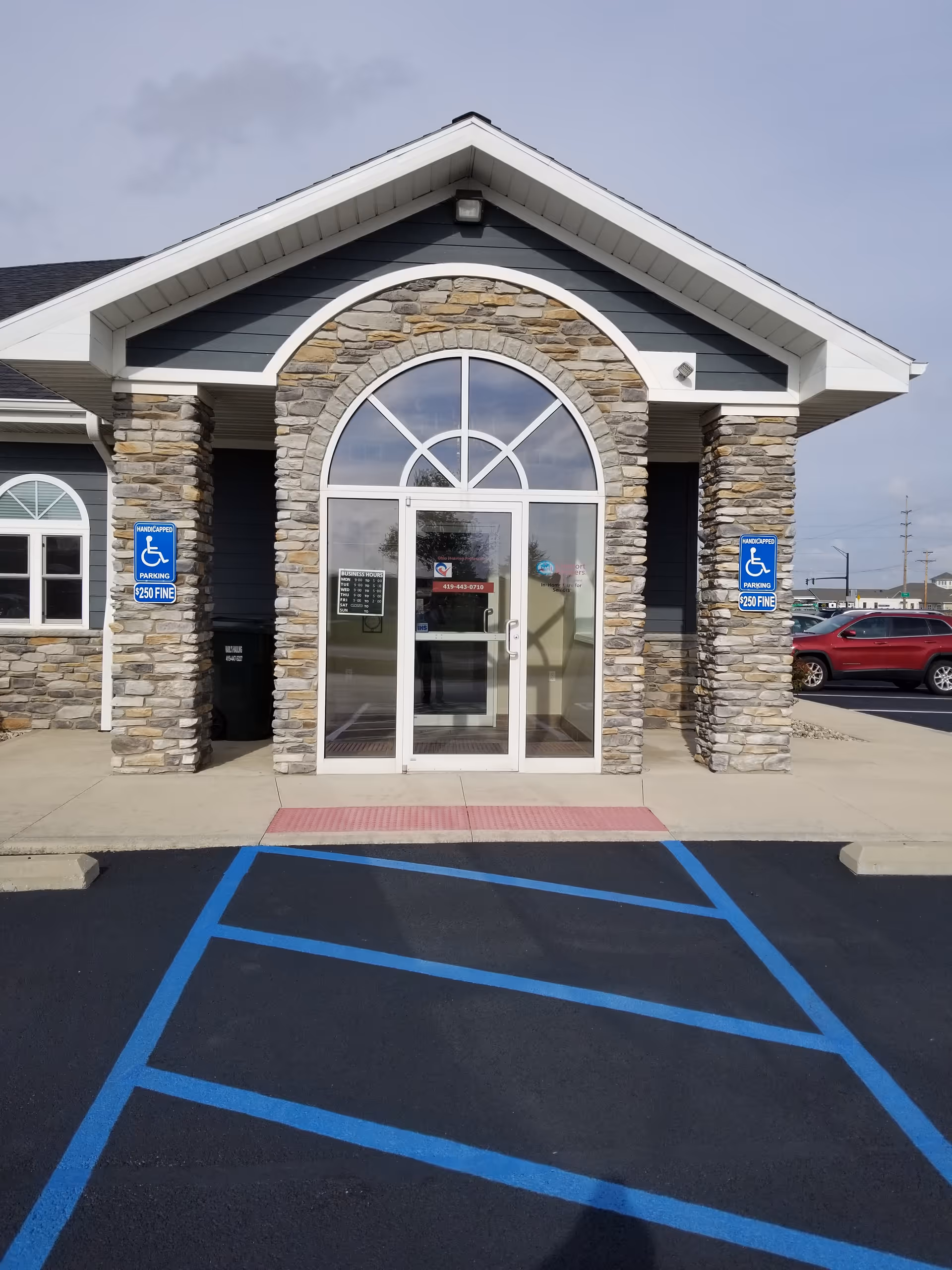 Front entrance of a stone-faced building with an arched window, wheelchair parking signs, and blue lines.