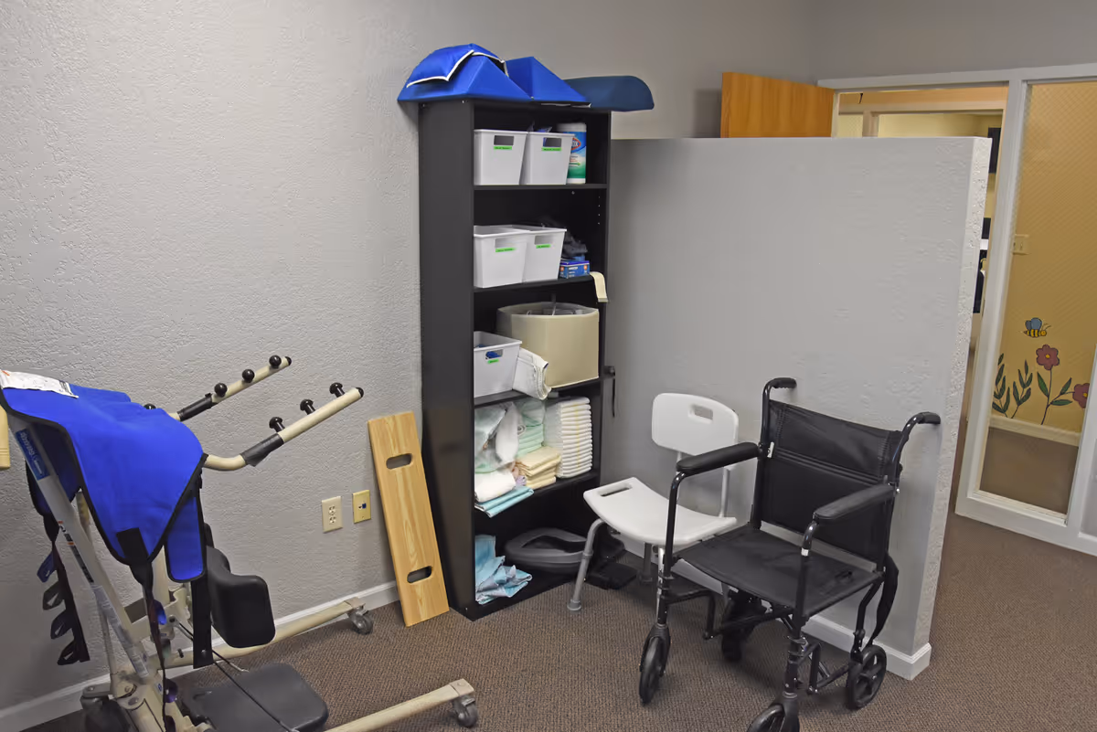 Storage corner with a tall shelf of medical supplies, a white chair, and a wheelchair.