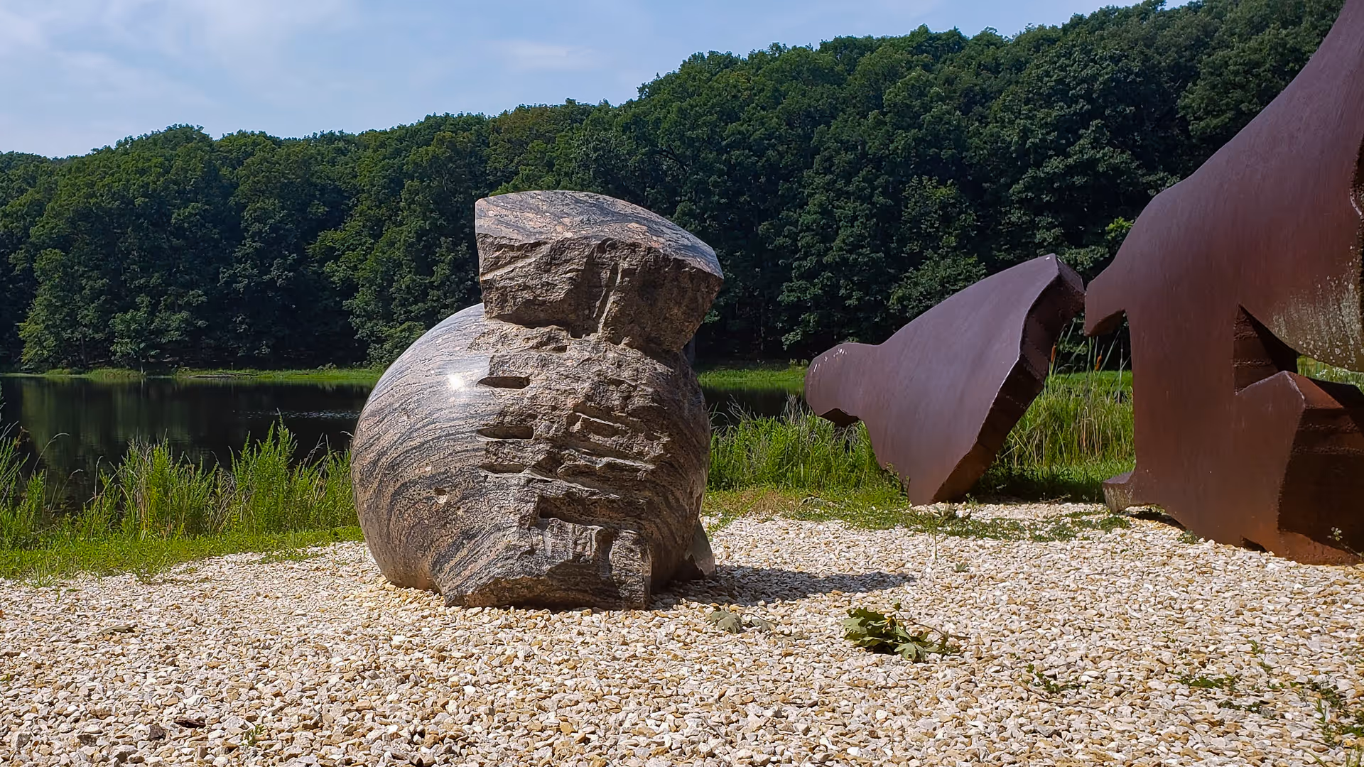 Outdoor park scene with a large stone sculpture and rusted metal art by a lake.