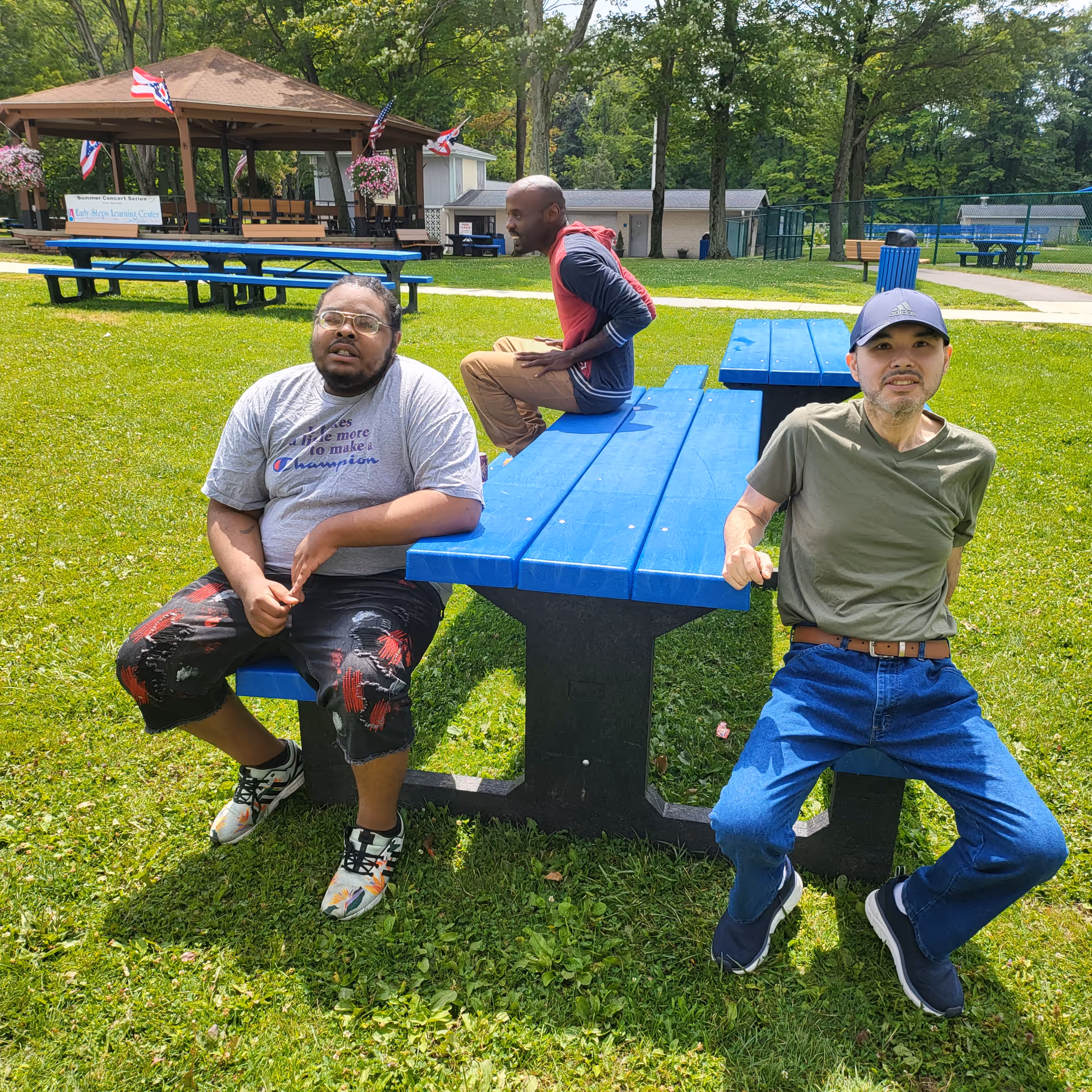 Three men sit at bright blue outdoor picnic tables in a sunny park.