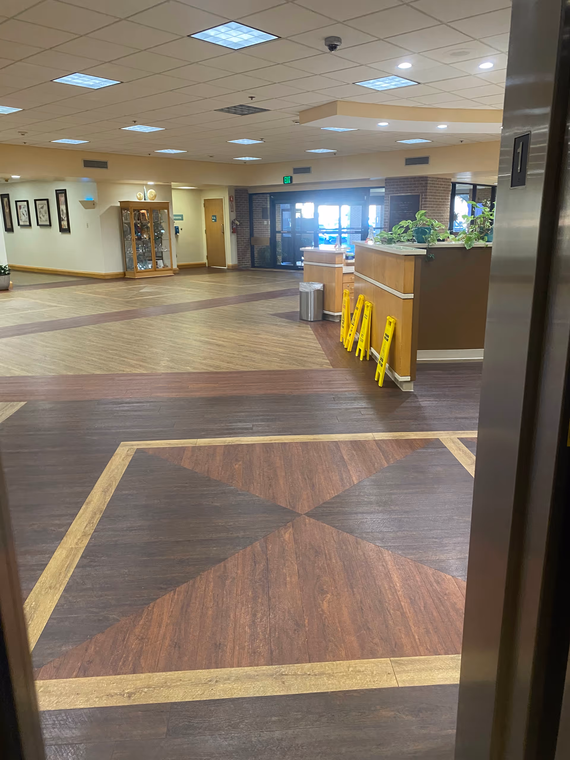Spacious indoor lobby with a reception desk, plants, and yellow wet floor signs.