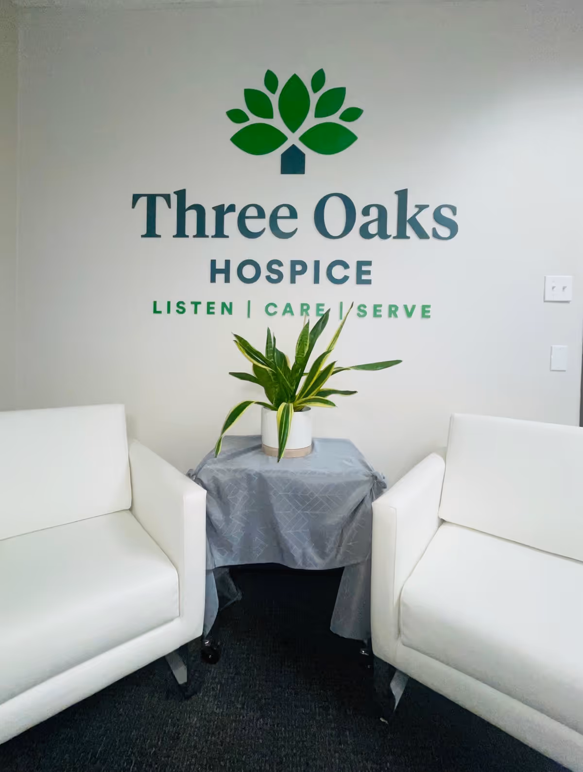 Lobby seating area with white chairs and a plant beside the Three Oaks Hospice logo.