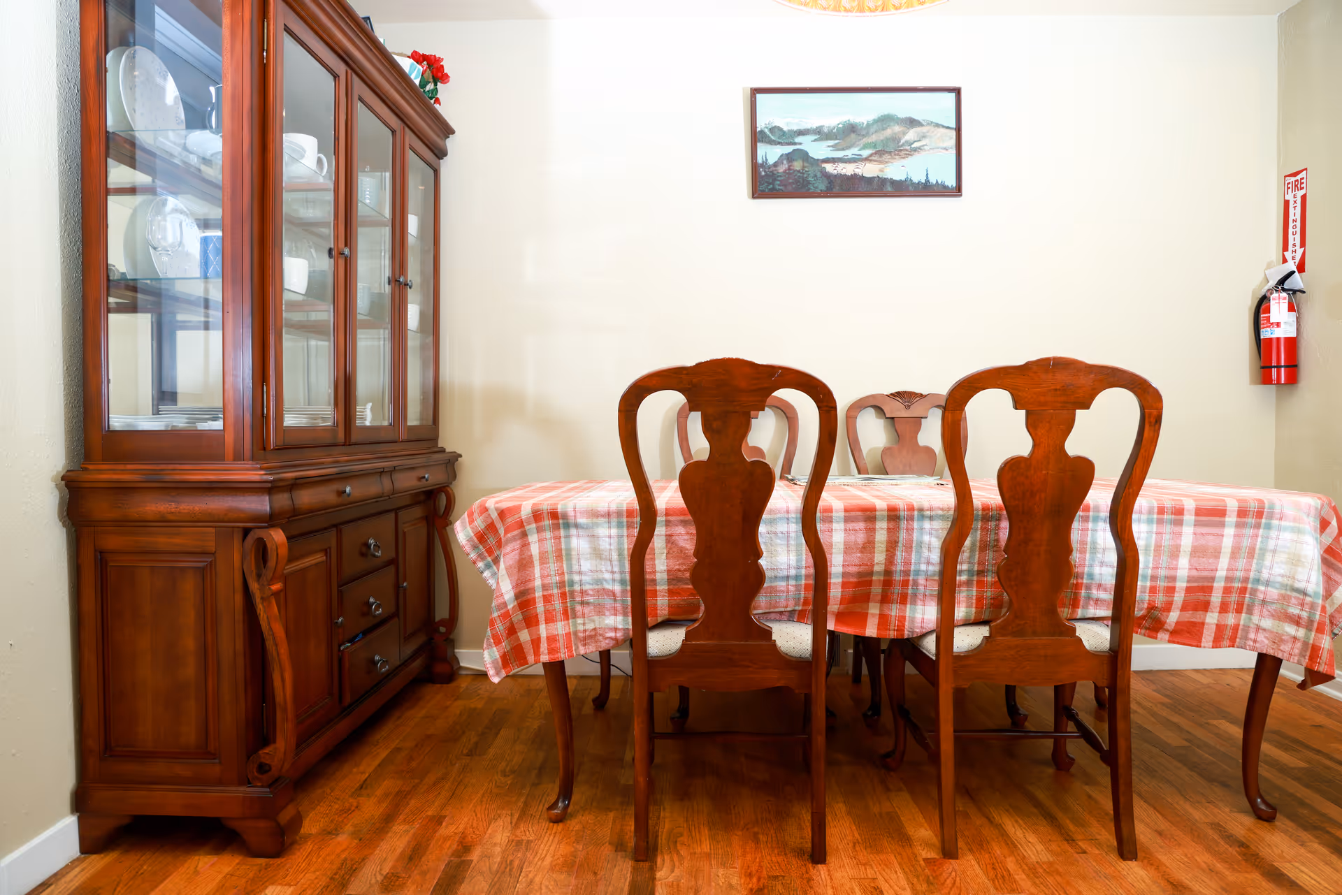 A wooden dining room with a table covered by a red plaid tablecloth and china cabinet.