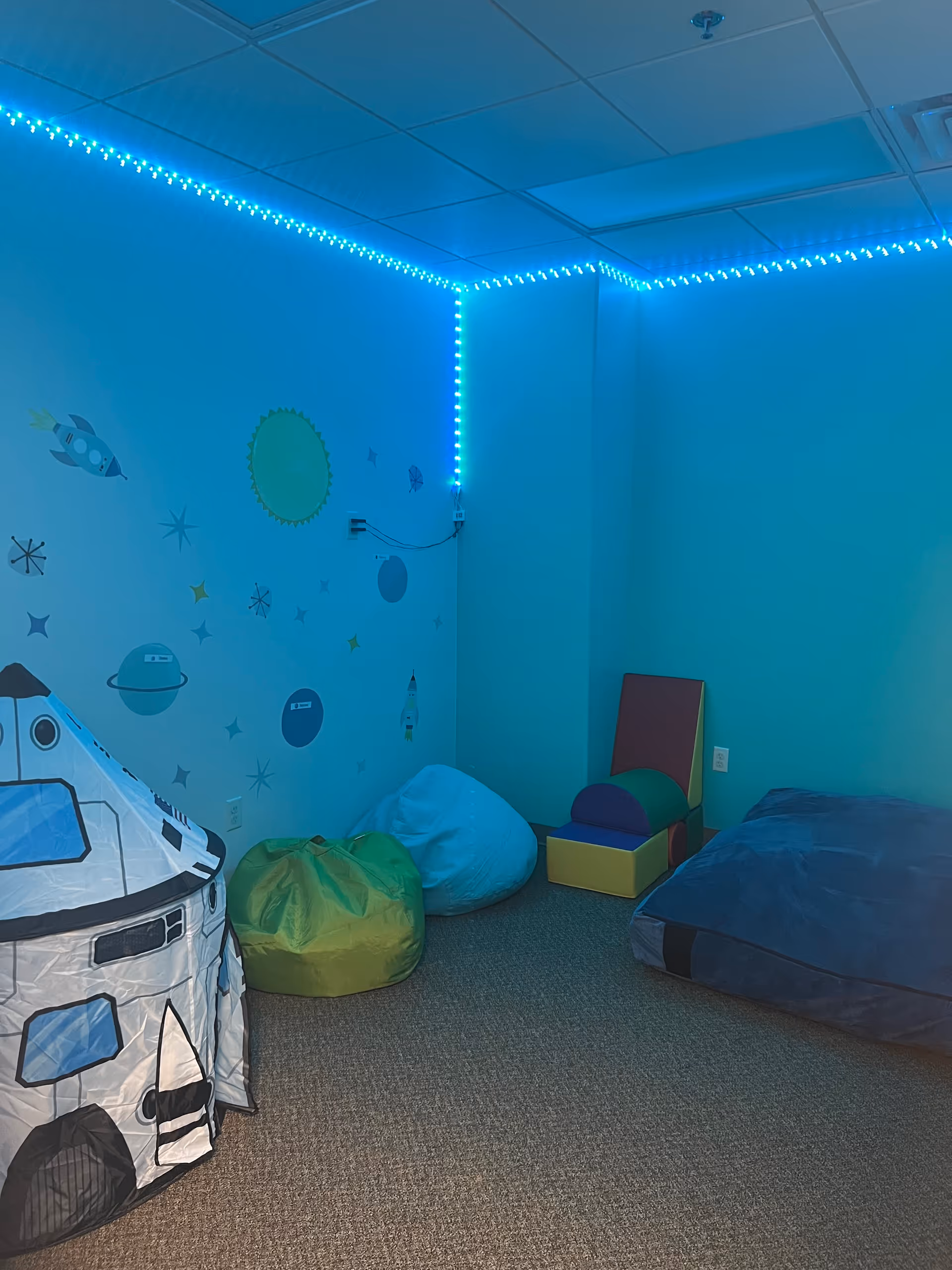 Blue-lit interior therapy room with space-themed decor, rocket tent, bean bags, and foam blocks.