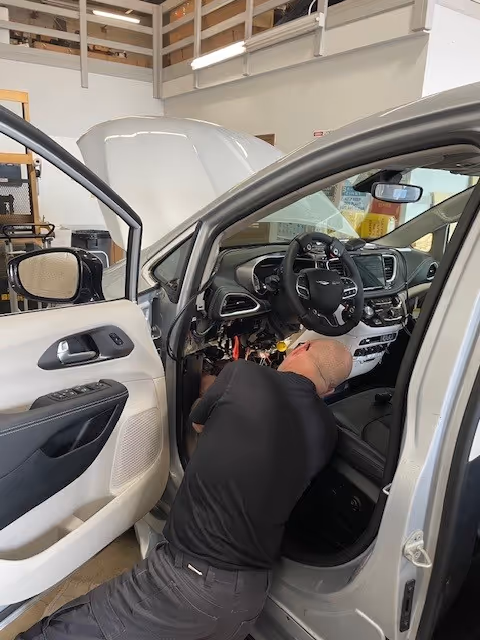 A technician works under the dashboard of a vehicle inside a service bay.