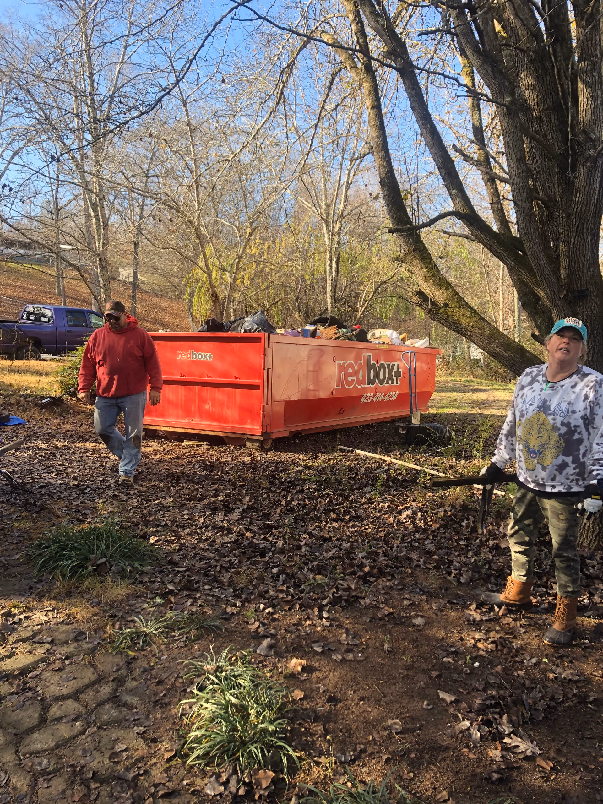 Two workers outdoors near a red dumpster, loading debris among leaf-covered ground.